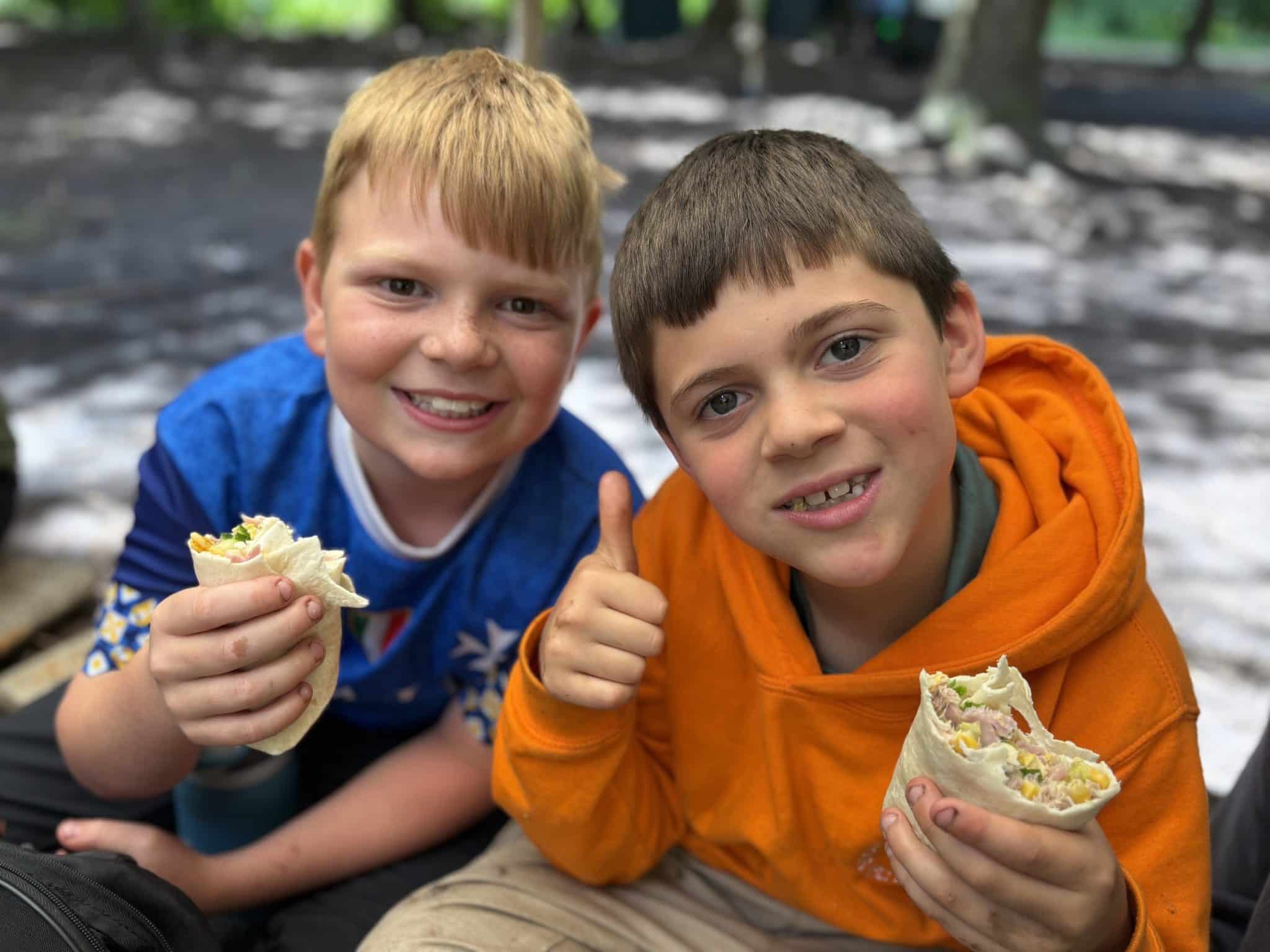 Two smiling boys sit outdoors, holding wraps filled with lettuce and other ingredients. One boy wears a blue shirt, and the other wears an orange hoodie and gives a thumbs-up. The background is blurred with trees and sunlight.