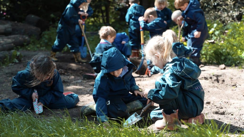 Children in blue raincoats play and dig with small tools in a muddy outdoor area surrounded by greenery at Cheltenham Prep School, engaged in group activities and exploring nature.