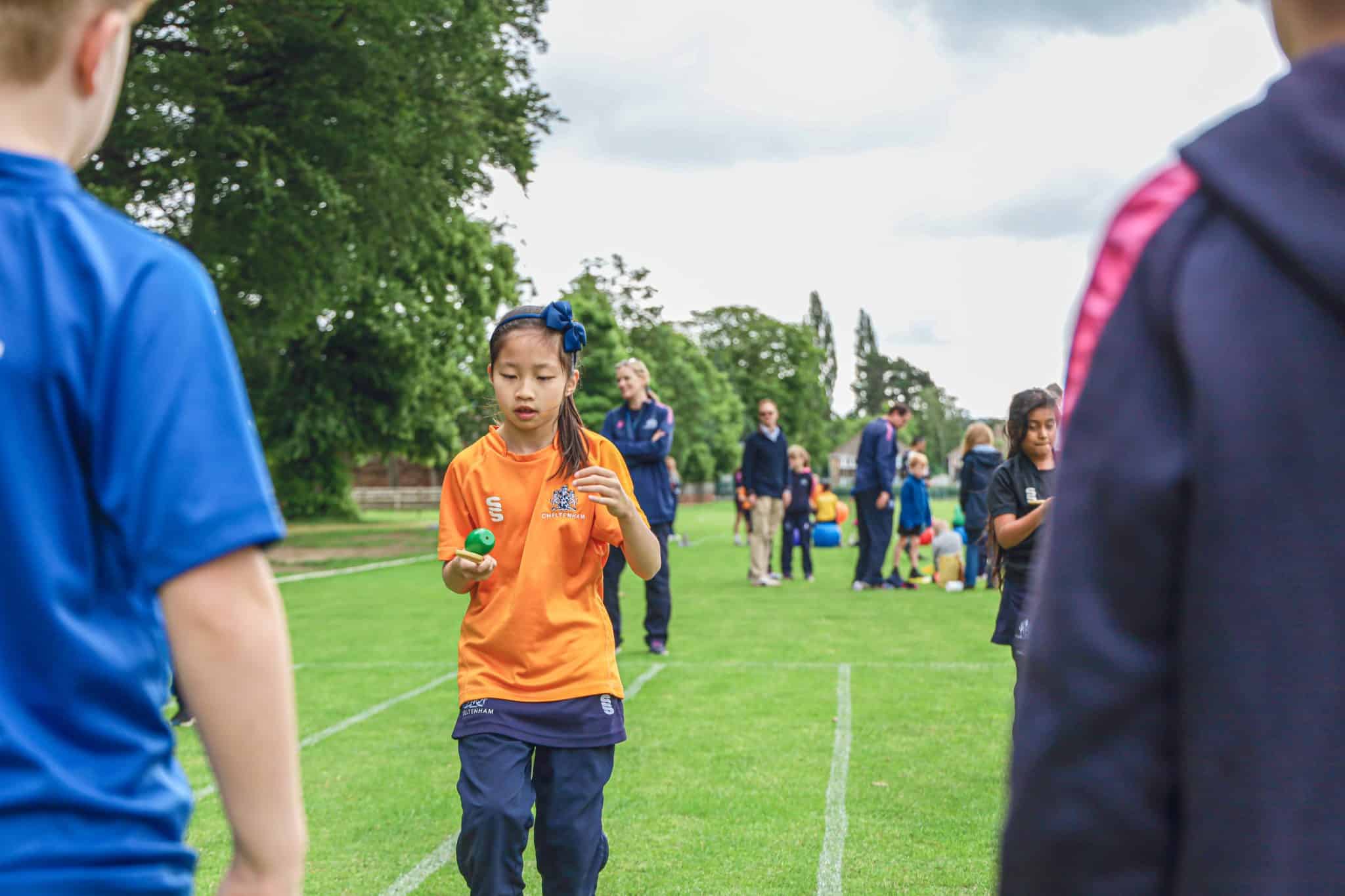 A young girl in an orange shirt balances an egg on a spoon during a race on a grassy sports field, with other children and adults in the background.