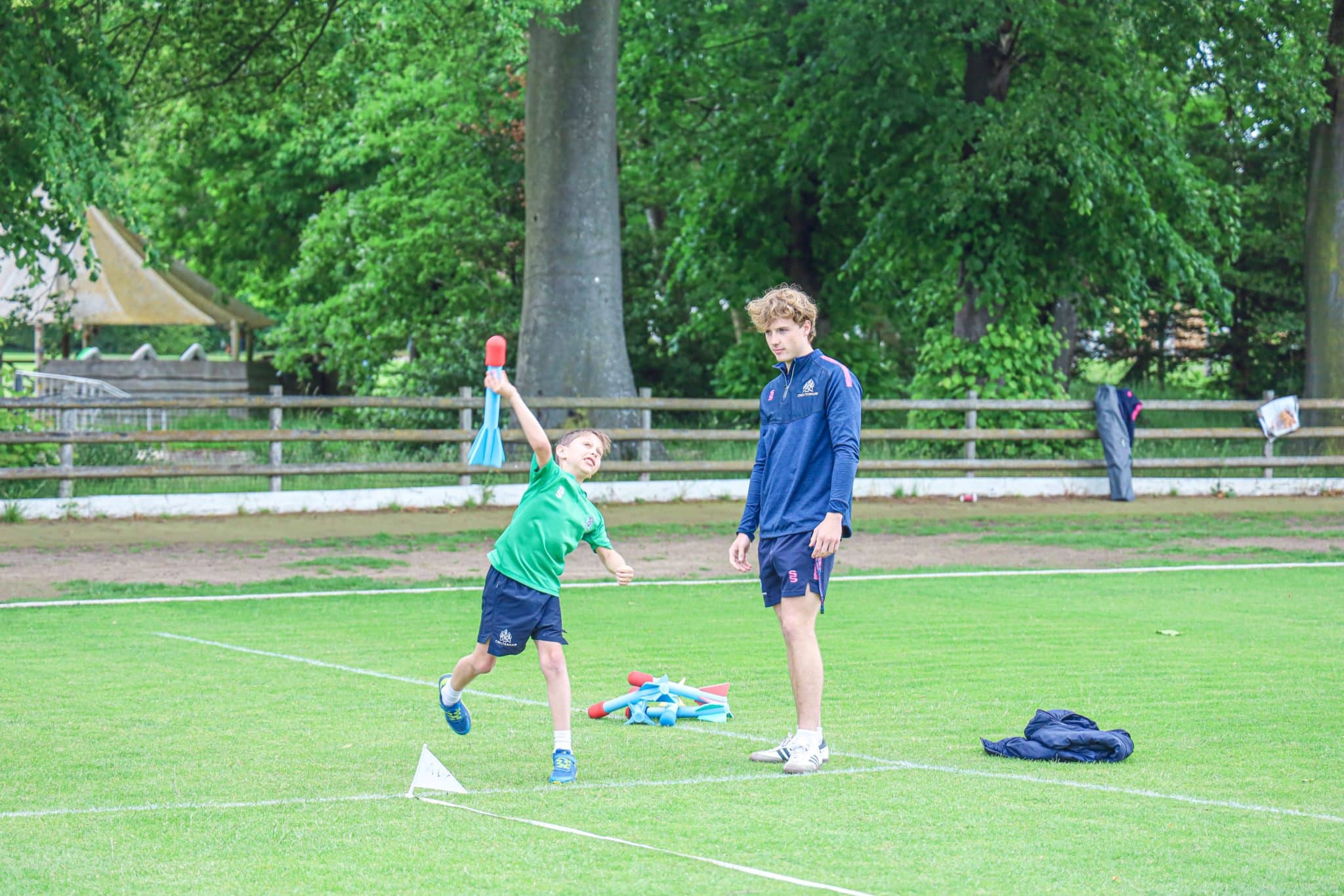 A young boy in a green shirt throws a small red object on a grassy field while an adult in sportswear watches. Trees and a wooden fence are in the background. Sports equipment lies on the ground nearby.
