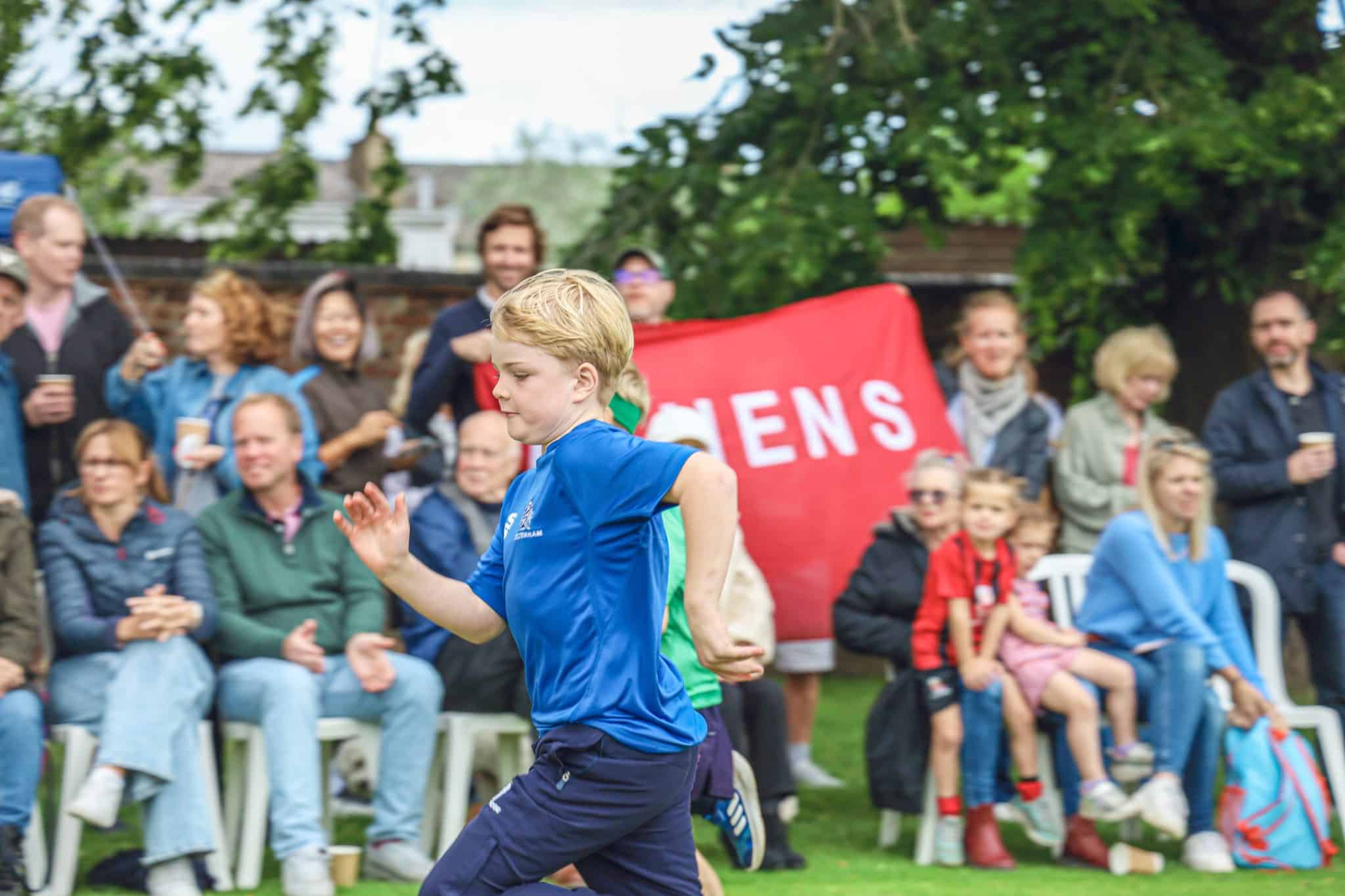 Two boys race on grass whilst a crowd of adults and children watch. A red banner with white letters is held up in the background. Trees and chairs are visible, suggesting an outdoor event or sports day.