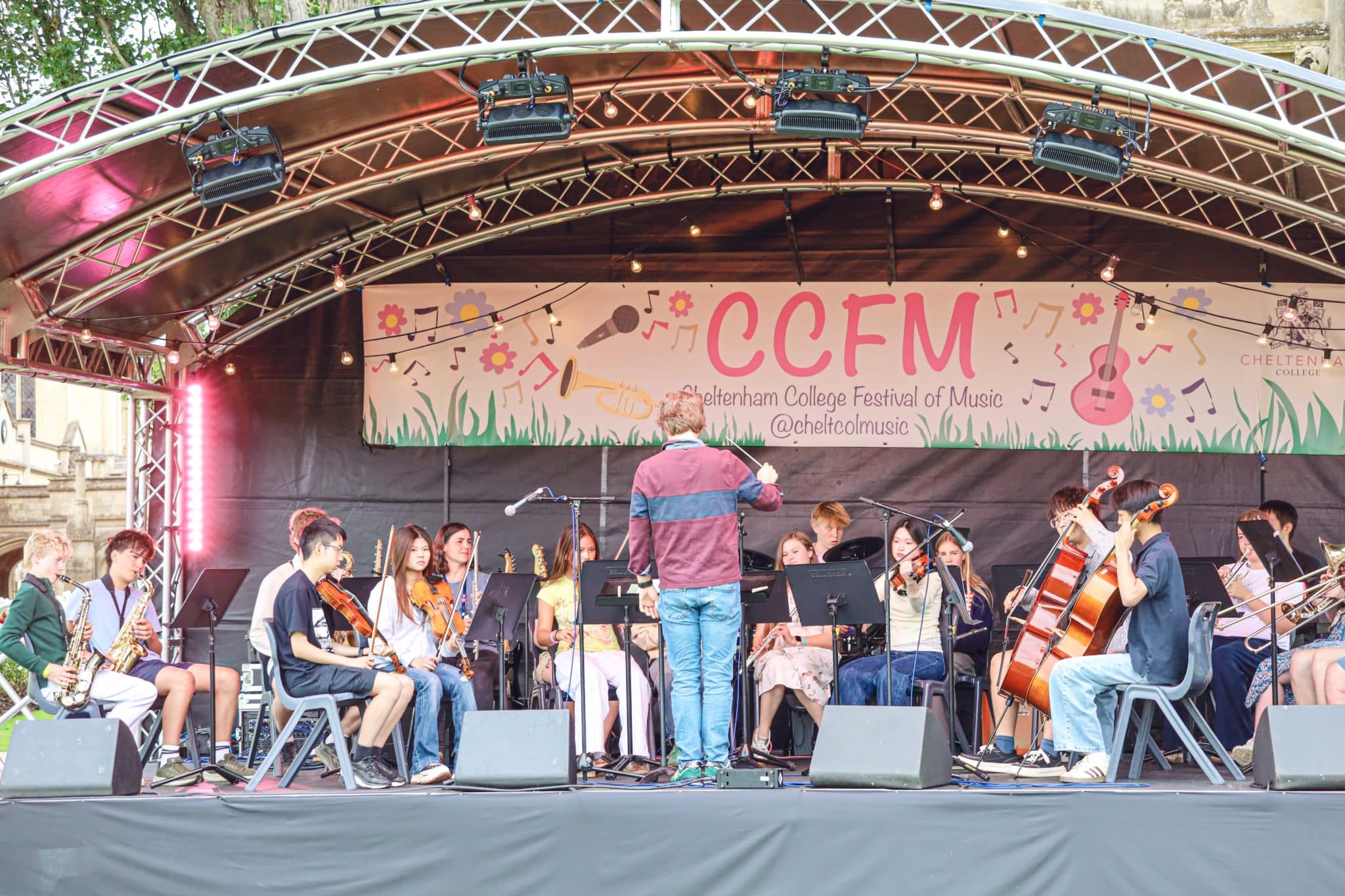 A conductor leads a youth orchestra performing on an outdoor stage under a banner reading CCFM, Chelterham College Festival of Music. The musicians play various instruments, including violin, cello, clarinet, and saxophone.