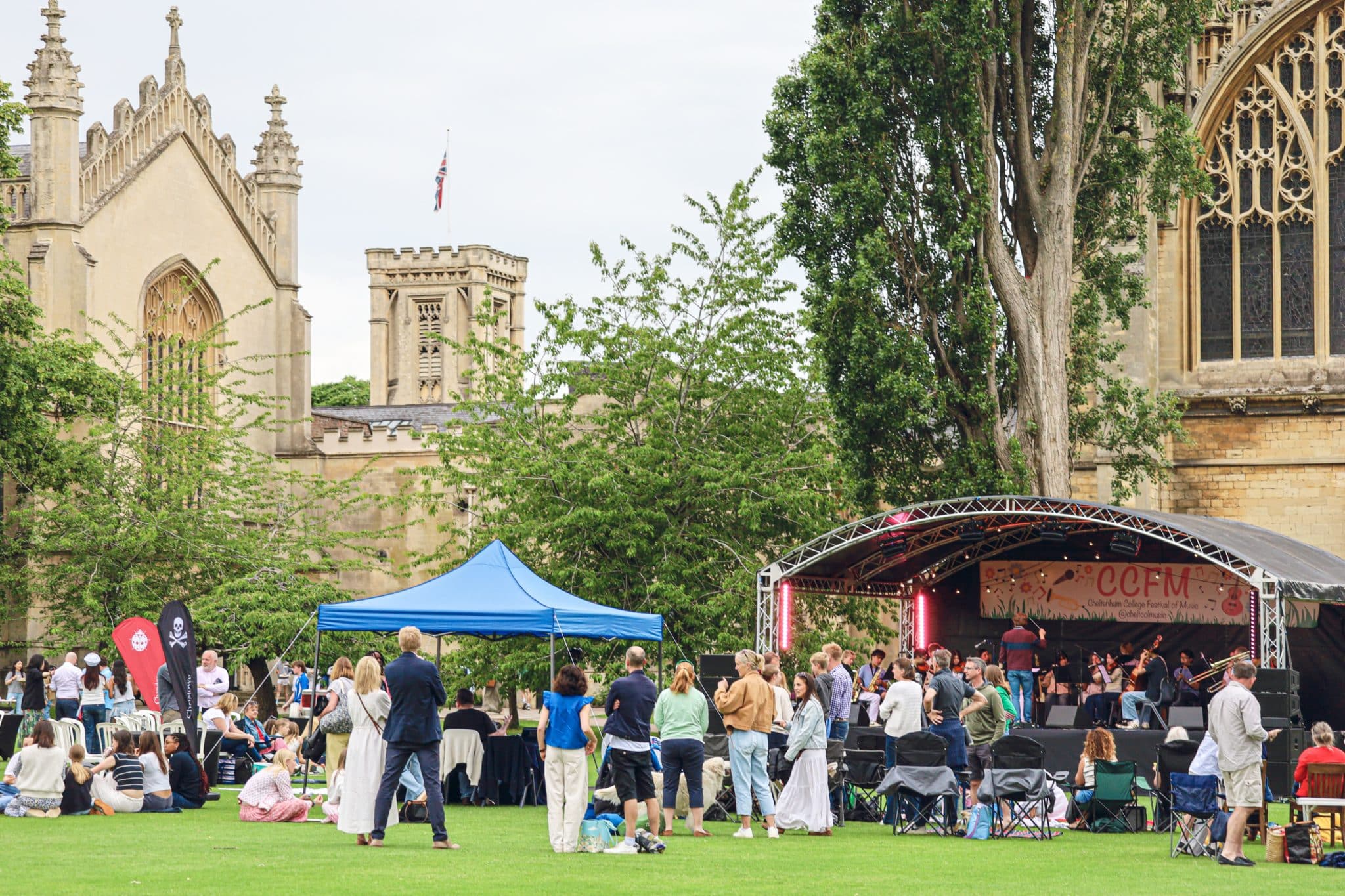 A crowd gathers on a grassy lawn near historic stone buildings to watch a live performance on an outdoor stage, with some people sitting and others standing near a blue marquee.