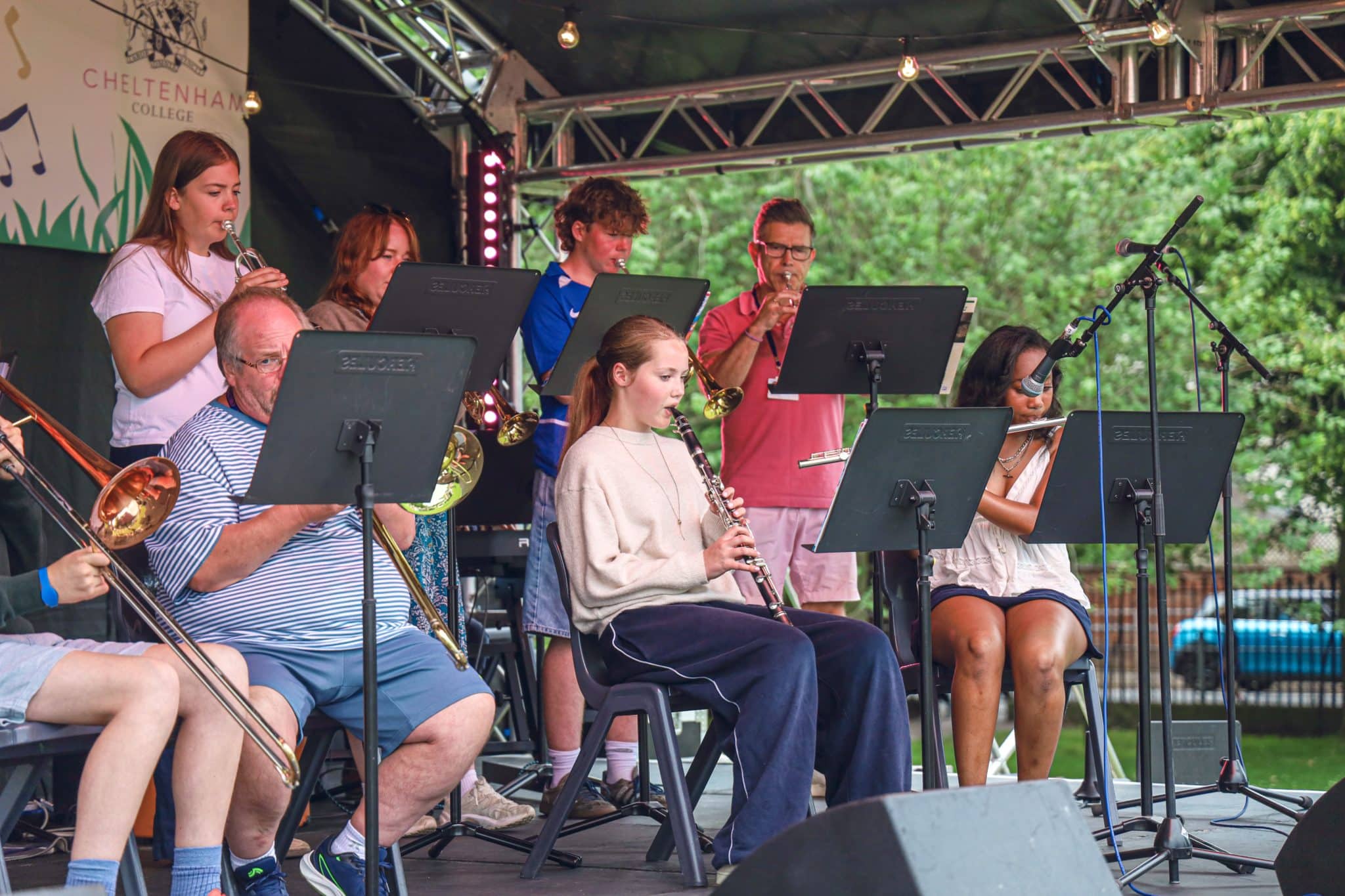 A group of people of various ages play wind instruments on an outdoor stage, reading music from stands, with trees and a banner in the background.