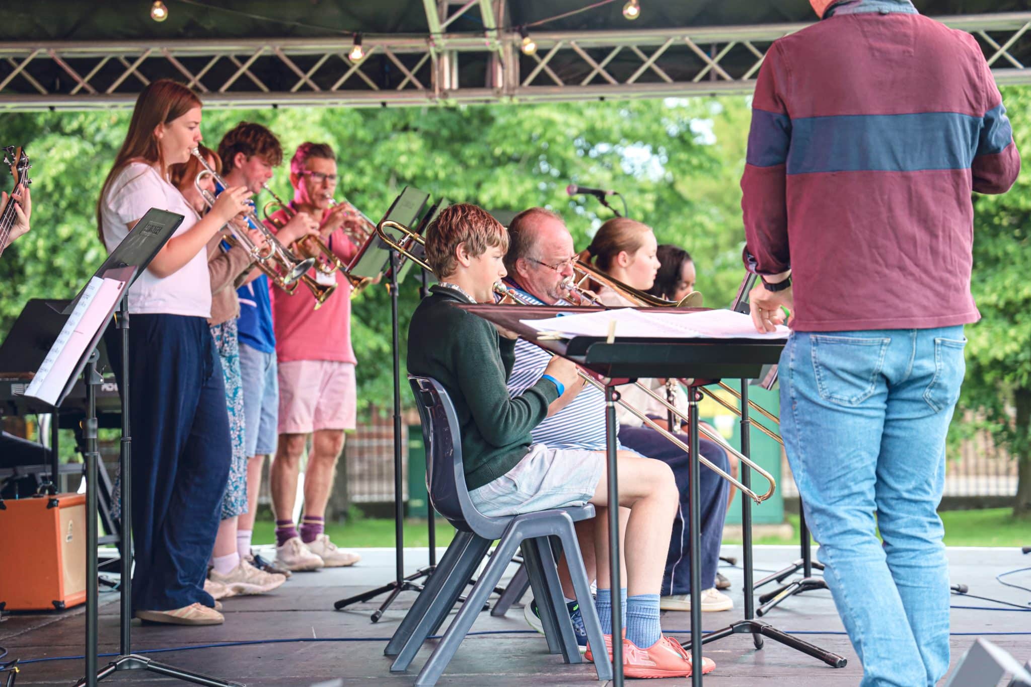 A group of musicians, some seated and some standing, play brass and wind instruments on an outdoor stage, guided by a conductor wearing a blue and maroon jacket. Trees are visible in the background.