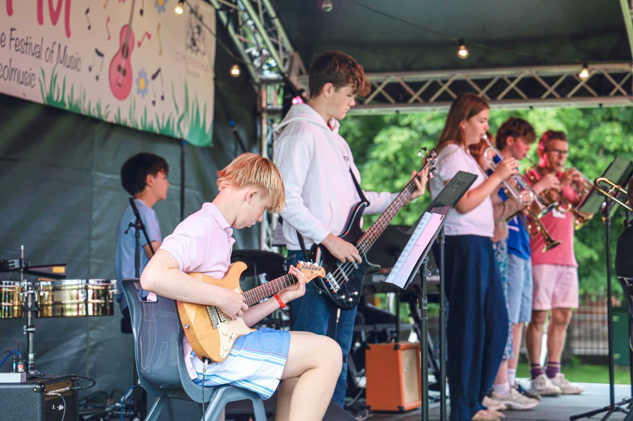 A group of young musicians perform on an outdoor stage, playing electric guitar, bass, keyboard, percussion, trumpet, and saxophone. They are dressed casually and focused on their instruments. Trees are visible in the background.