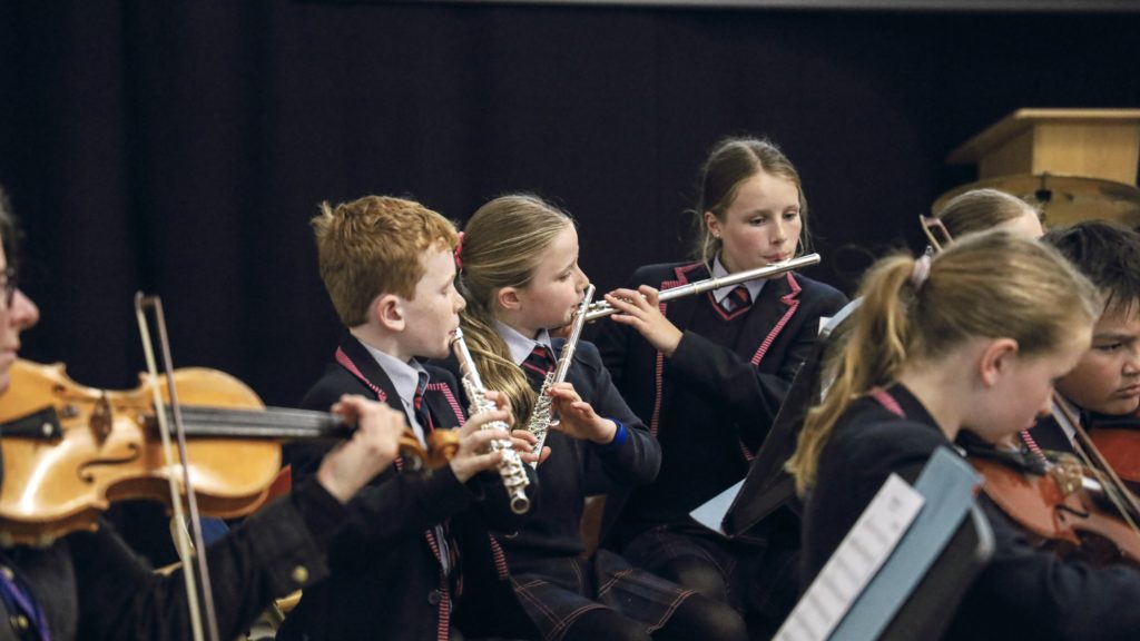 A group of Cheltenham Prep School children in uniforms play instruments, including violins and flutes, during a music performance.