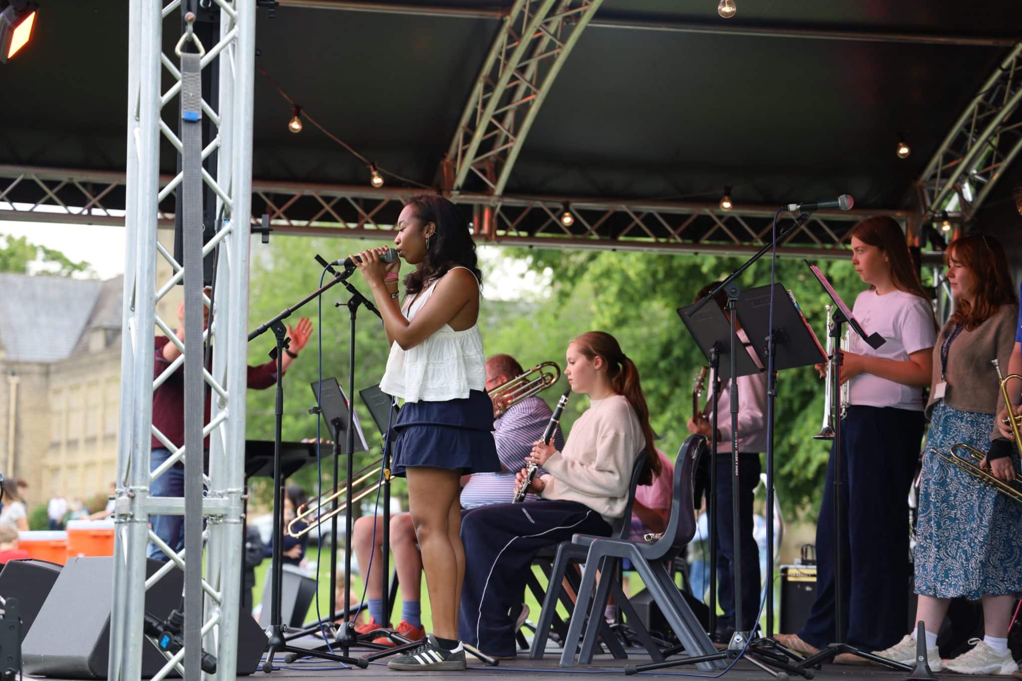 A young woman sings into a microphone on an outdoor stage with several musicians playing instruments like trombone, clarinet, and saxophone behind her. The stage is covered and set up in a park with trees in the background.