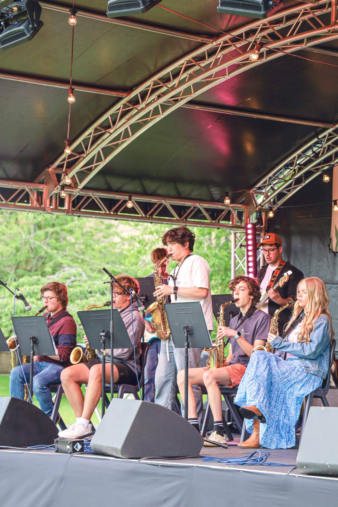 A group of young musicians perform on an outdoor stage, playing various instruments including saxophones and a guitar. They are seated under a canopy with music stands in front of them. Trees are visible in the background.