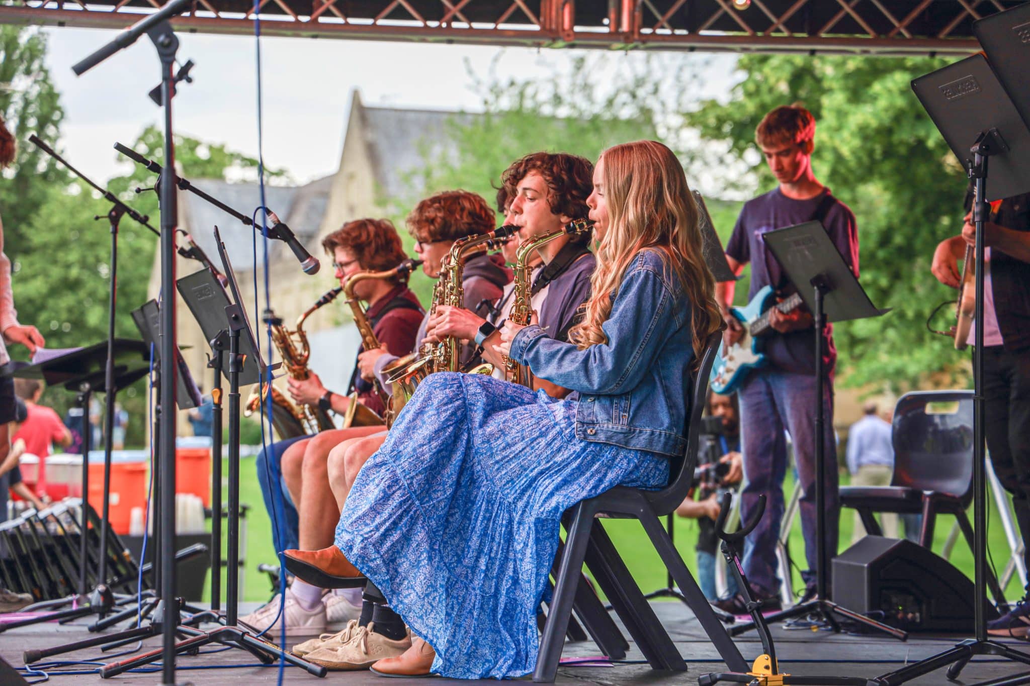 A group of young musicians perform on an outdoor stage, playing saxophones and electric guitar. A girl in a blue dress and denim jacket sits in the foreground, focused on her instrument. Trees and a building are in the background.