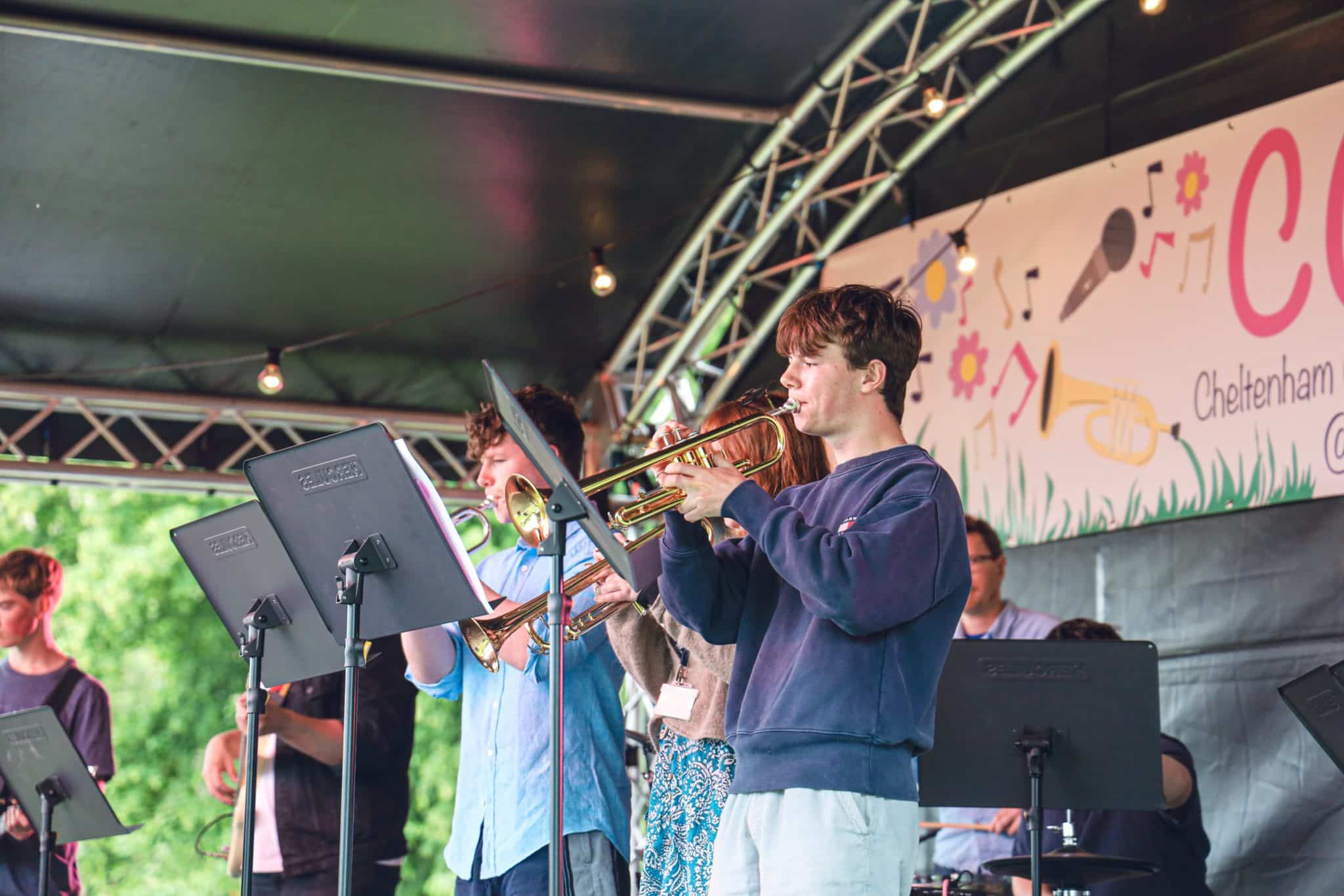 A group of young musicians play brass instruments on an outdoor stage, with a festival banner and fairy lights overhead. Music stands are in front of them, and greenery is visible in the background.