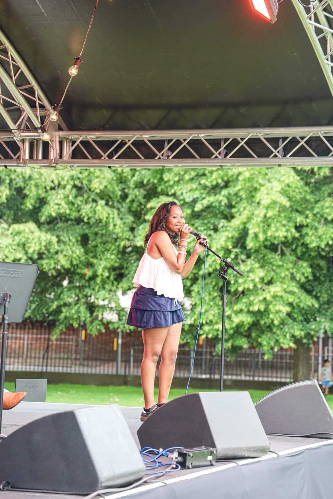 A woman in a white top and dark skirt sings into a microphone on an outdoor stage, surrounded by loudspeakers and trees in the background.