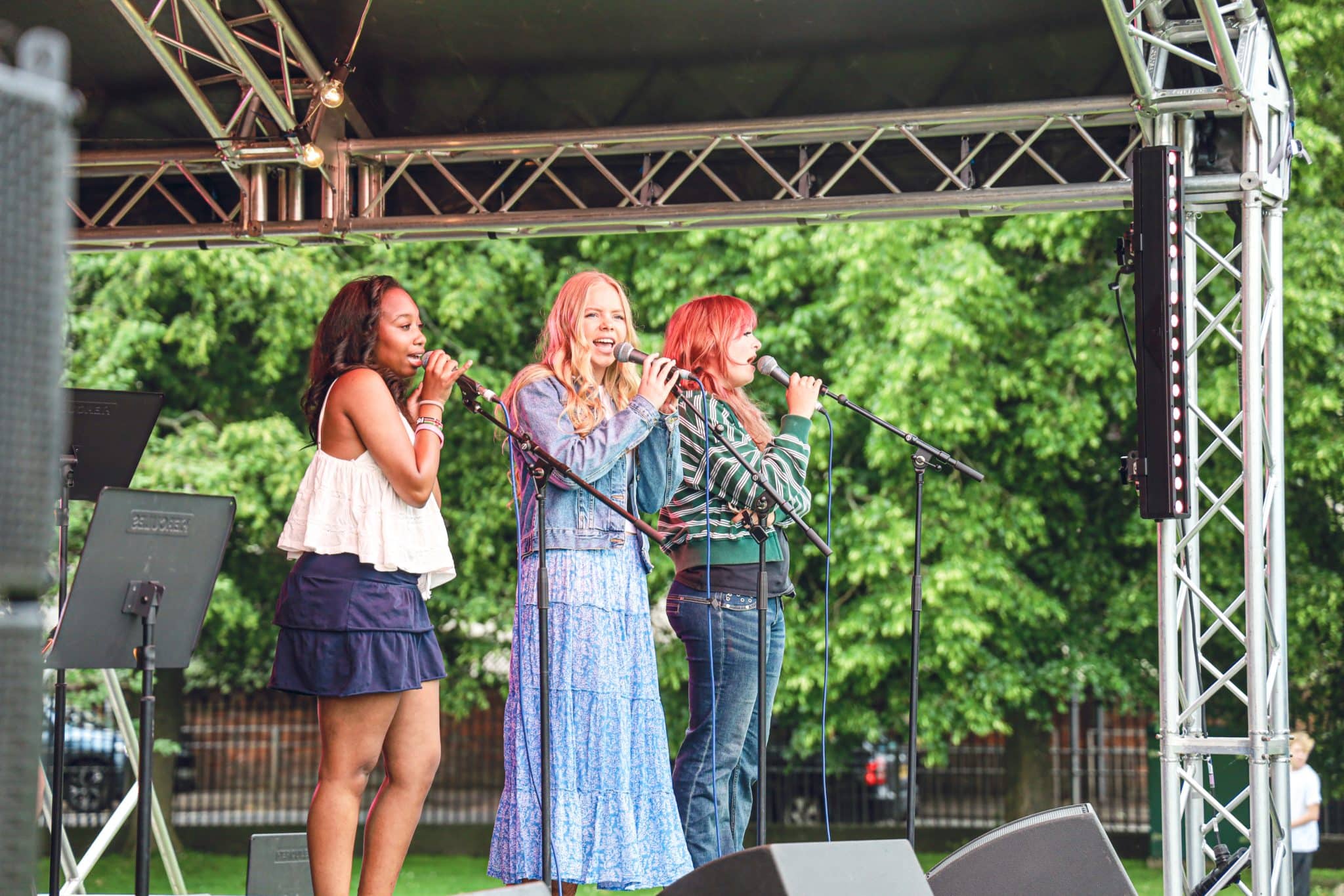Three women sing into microphones on an outdoor stage, standing in front of green trees and metal scaffolding. They appear to be performing at a live event or concert, smiling as they sing together.