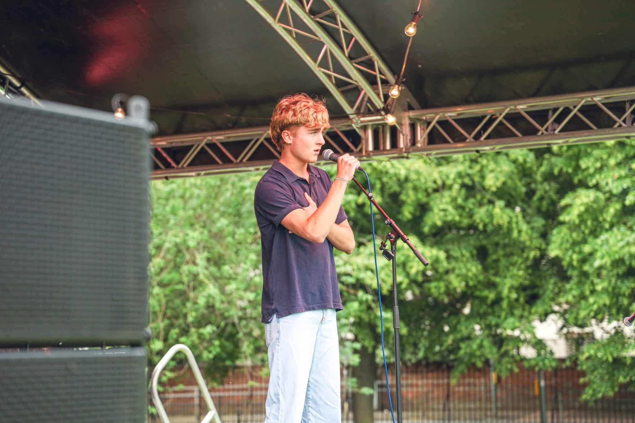 A person with short, wavy blonde hair stands on an outdoor stage, speaking or singing into a microphone. They wear a navy shirt and light blue trousers, with green trees and stage lights in the background.