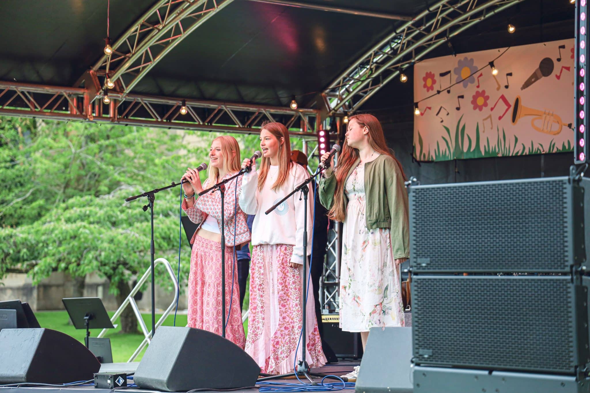 Three young women sing into microphones on an outdoor stage decorated with colourful artwork. They stand close together, each holding a microphone, surrounded by speakers and stage equipment, with greenery visible in the background.