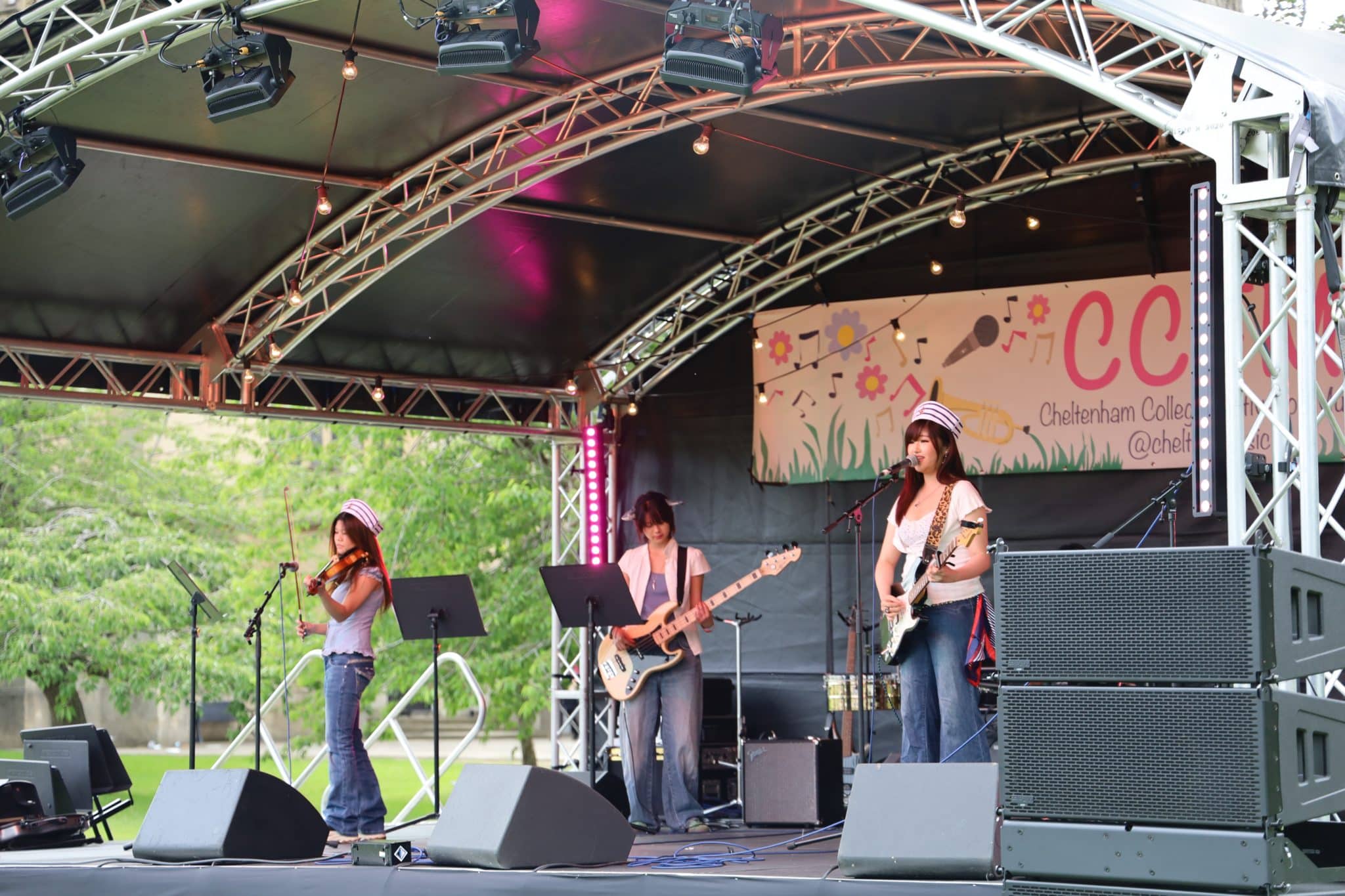 A three-member band performs on an outdoor stage, with one playing violin, one on bass guitar, and one on electric guitar and singing. They wear casual clothes and hats. A colourful event banner hangs behind them.