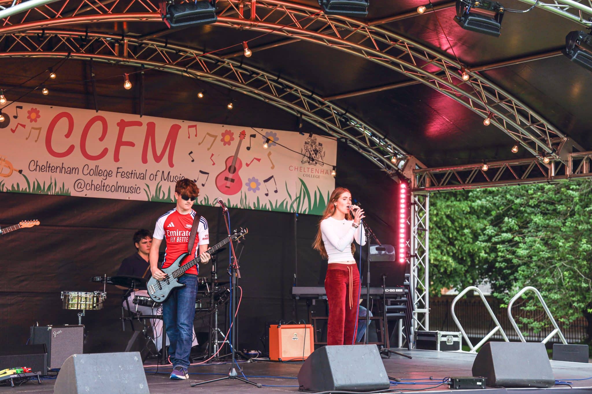 A band performs on an outdoor stage at the Cheltenham College Festival of Music. The female singer holds a microphone whilst other band members play instruments under a lit, covered stage.