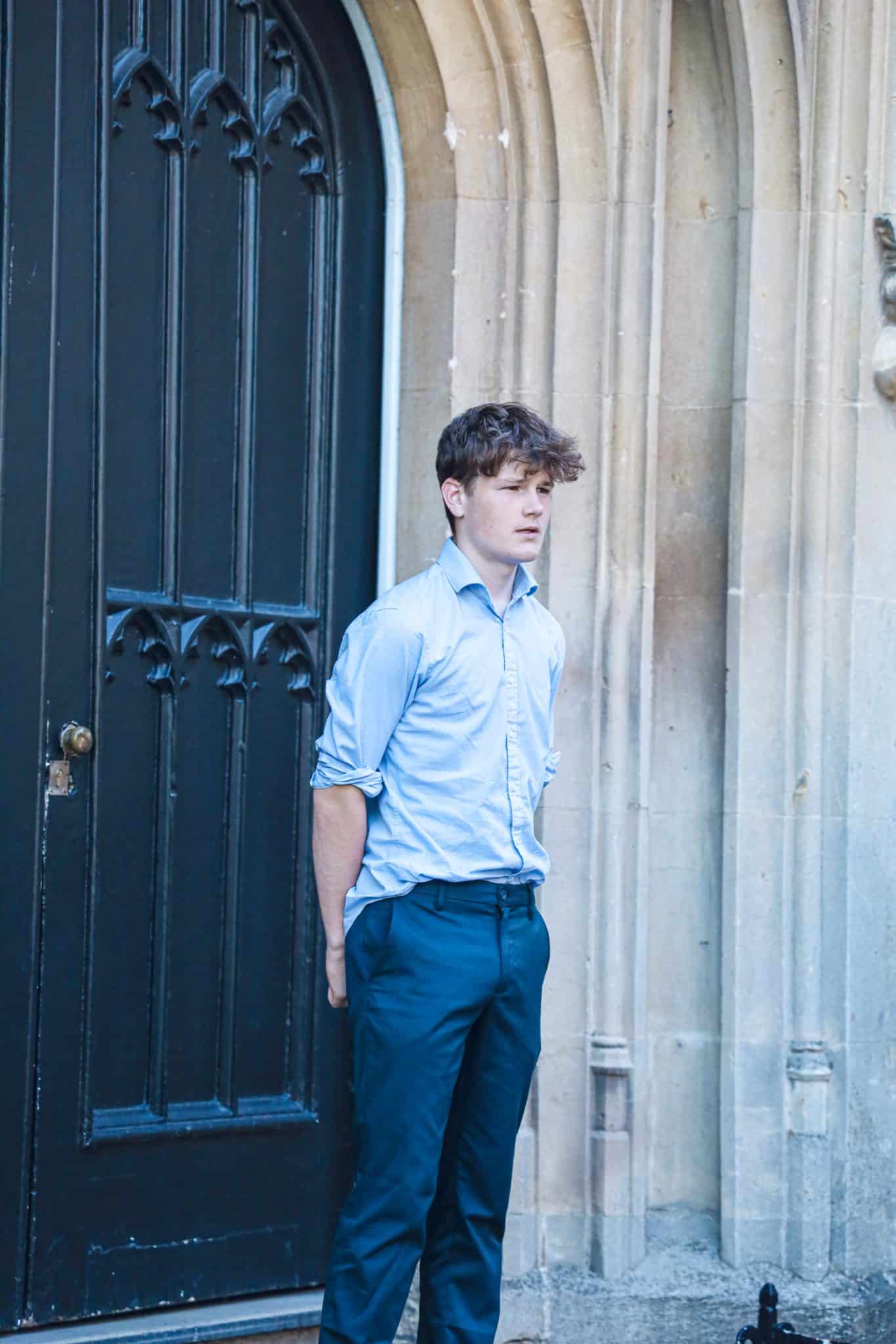A young man in a light blue shirt and dark blue trousers stands with his hands behind his back in front of a large, ornate black door set in a stone building.