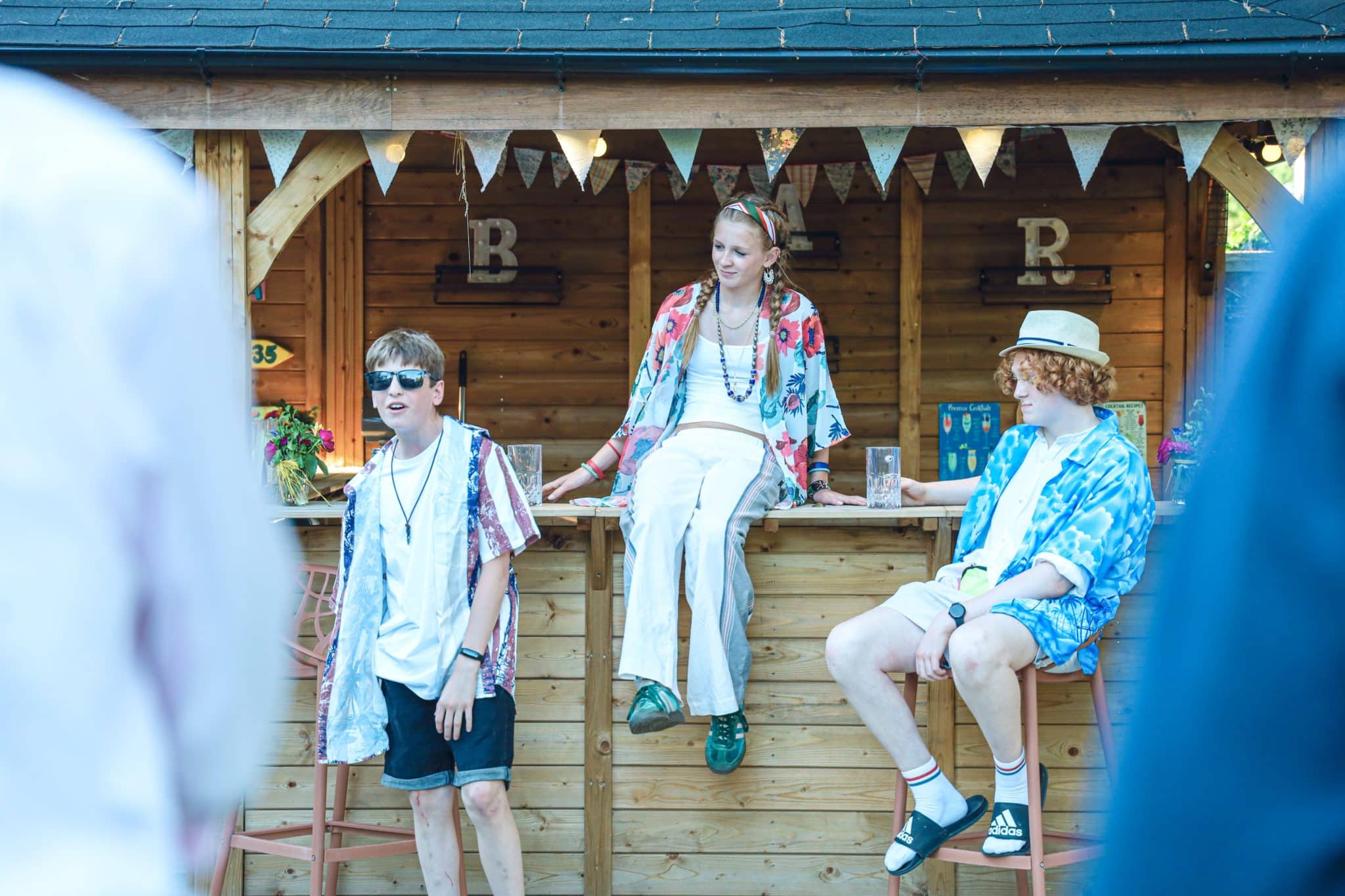 Three young people sit and stand by a wooden outdoor bar decorated with bunting. They wear summer clothes and sunglasses, with casual, relaxed expressions. The area is sunlit and festive.