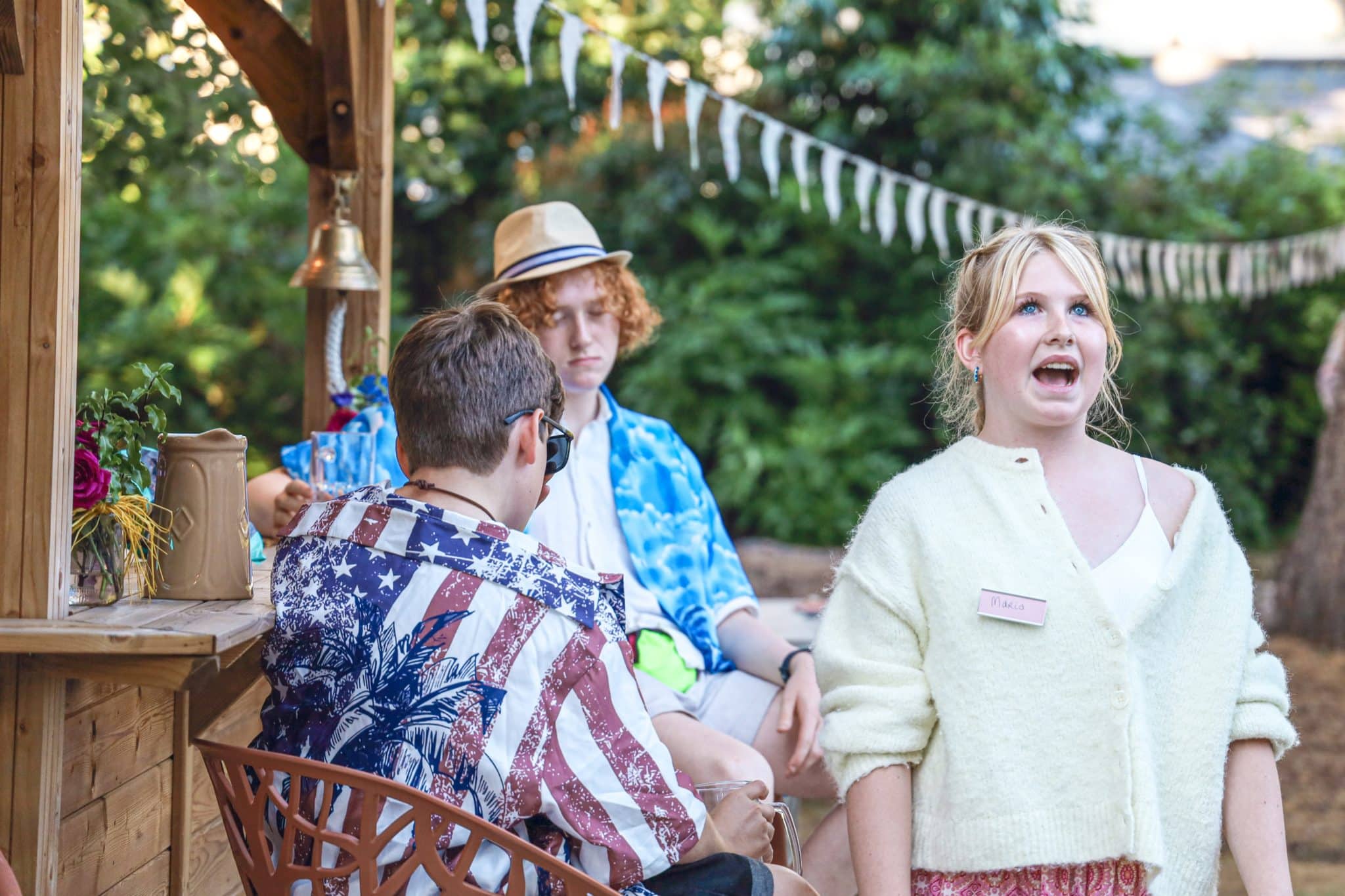 A woman speaking outdoors wears a cream cardigan and name badge, while two men sit nearby under a wooden canopy, one in a blue shirt and hat. String bunting decorates the background with greenery visible.