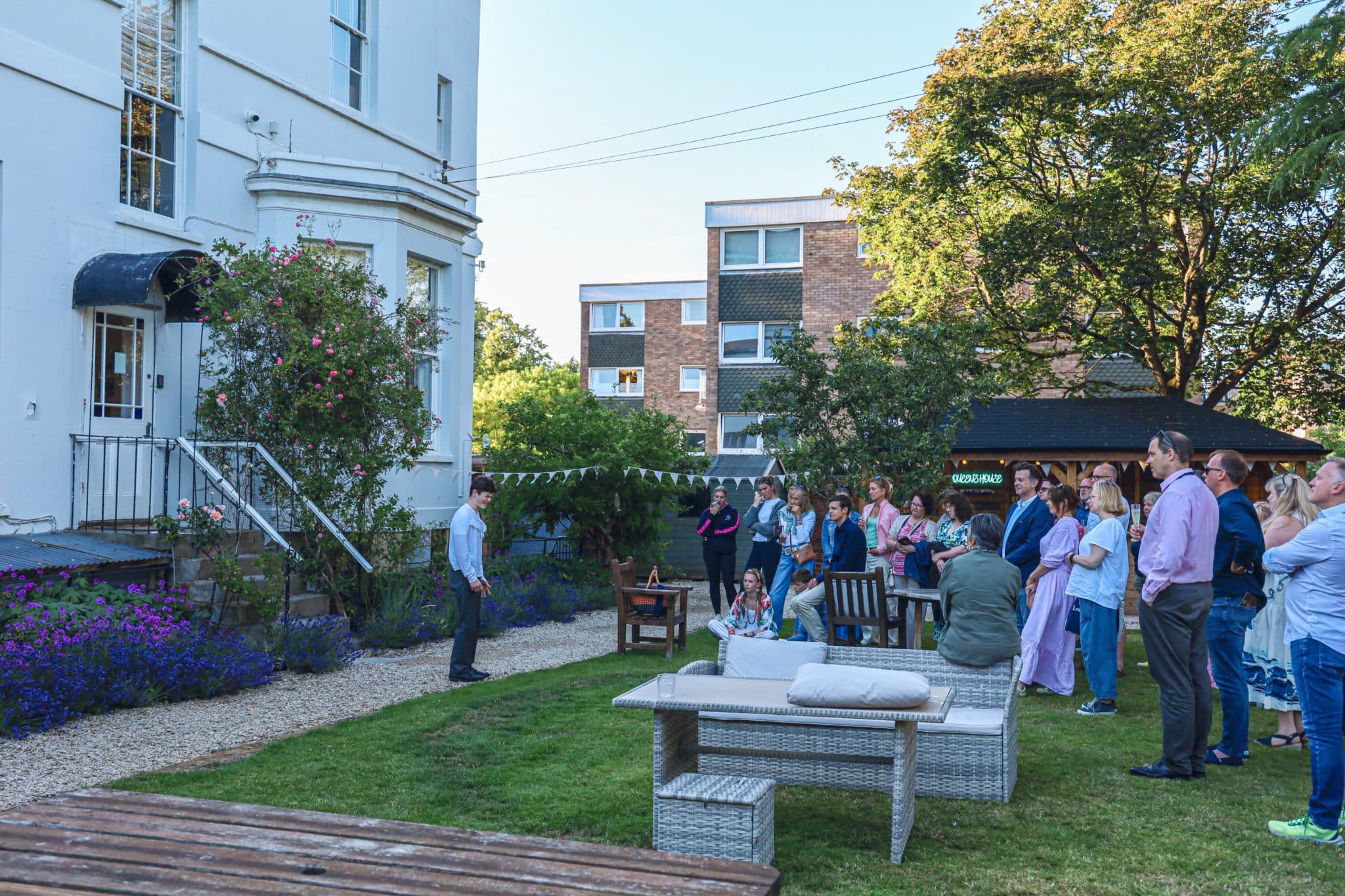 A group of people stands on a grassy lawn, listening to a man speaking near the steps of a white building. There are trees, benches, and nearby block of flats in the background.