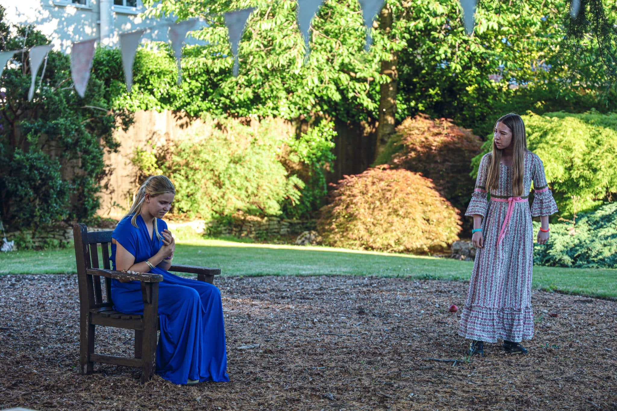 Two women in period costumes are in an outdoor setting. One in a blue dress sits on a wooden chair looking down, while the other in a patterned dress stands, appearing to speak to her. Trees and bunting are in the background.