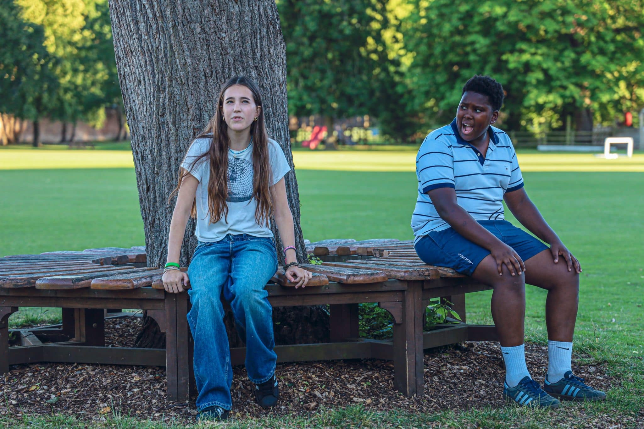 Two teenagers sit on a wooden bench around a tree in a park. One looks up with a thoughtful expression, whilst the other looks at them, appearing surprised. Lush green grass and trees fill the background.