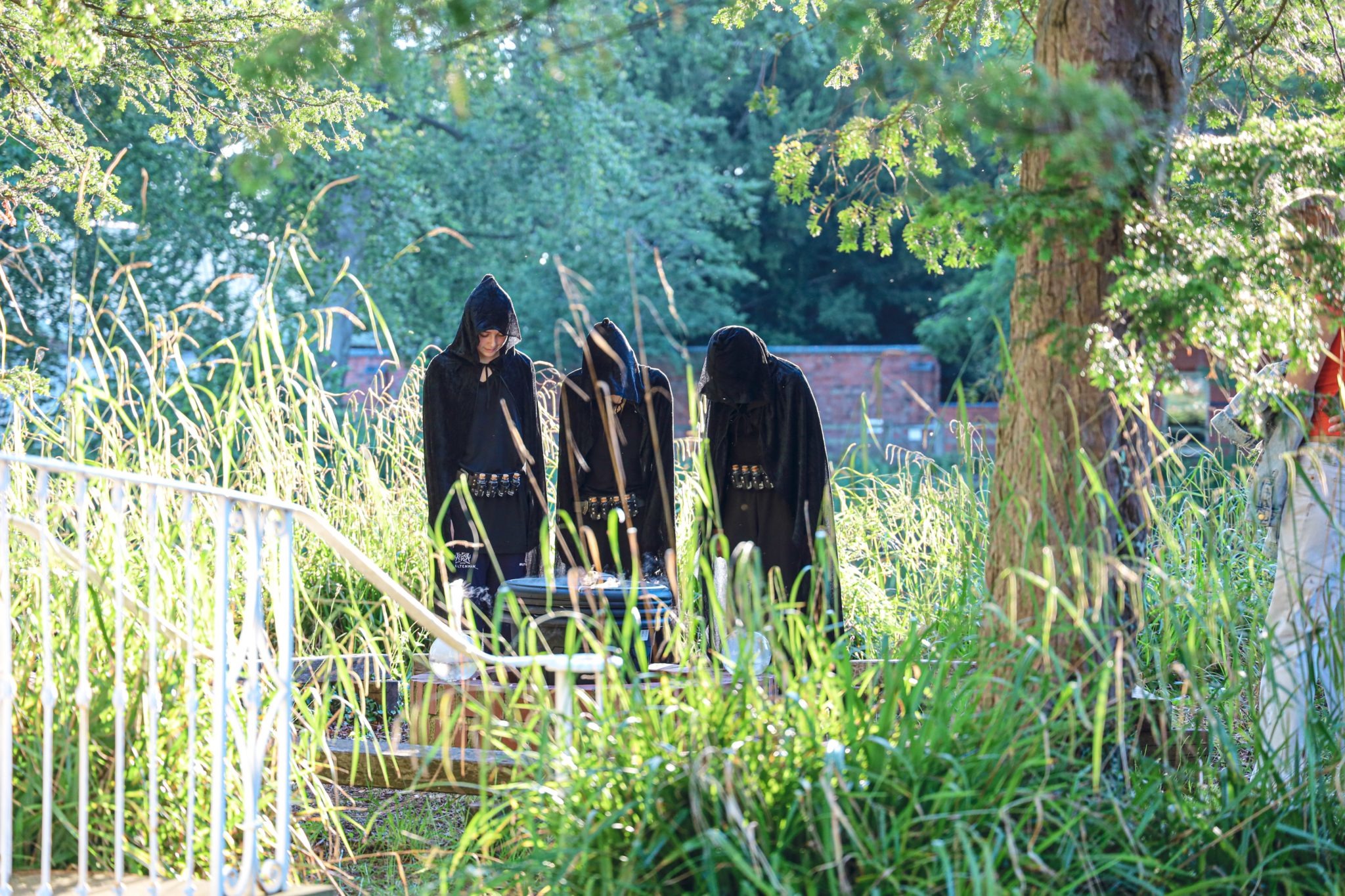 Three people in black hooded cloaks stand closely together in tall grass and sunlight, with trees and greenery around them. They appear to be participating in an outdoor, possibly ceremonial or theatrical event.