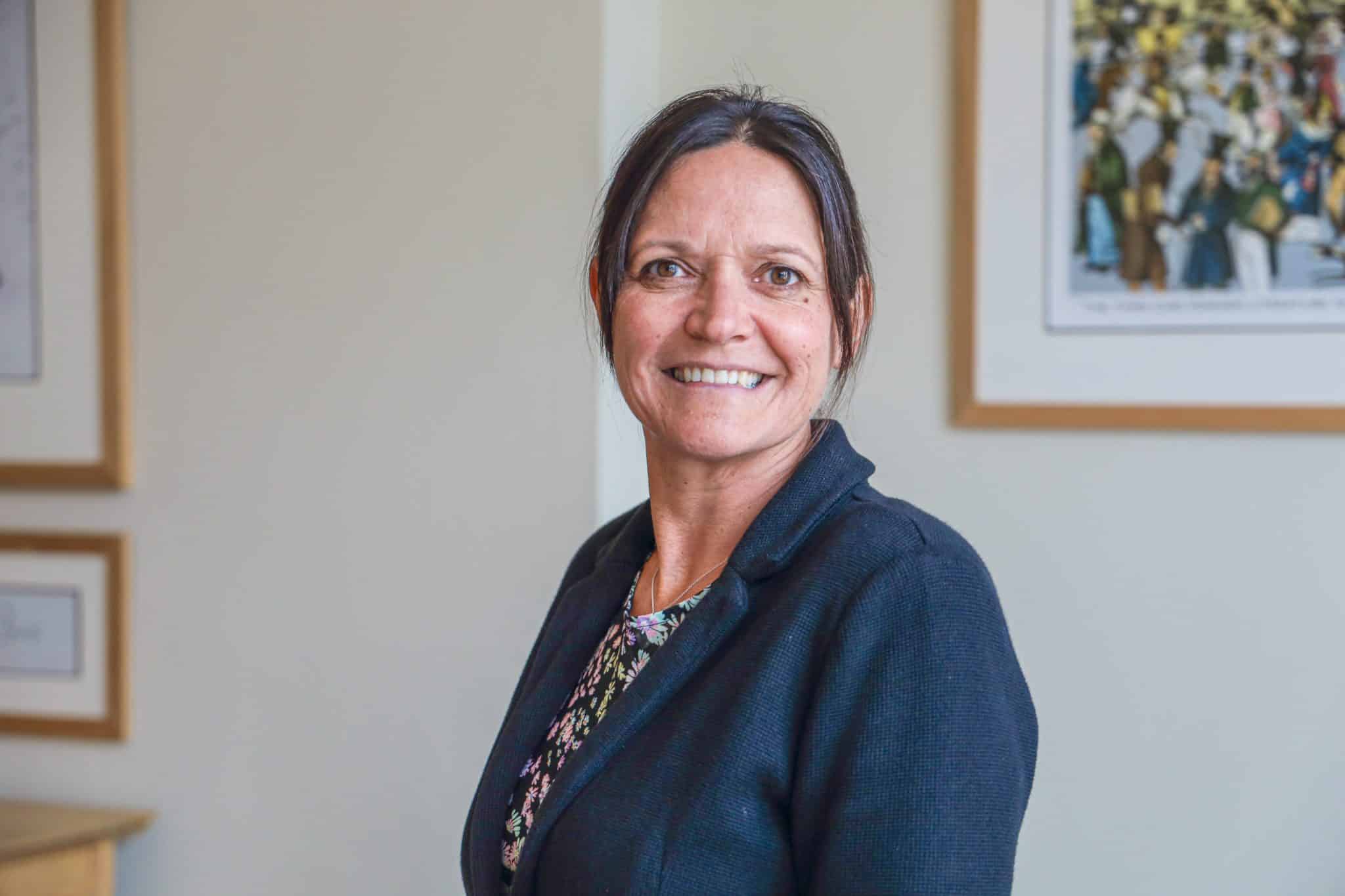 A woman with dark hair tied back, wearing a dark blazer and floral top, smiles whilst standing indoors in front of framed artwork on a light-coloured wall, reflecting the welcoming atmosphere of Cheltenham College admissions.