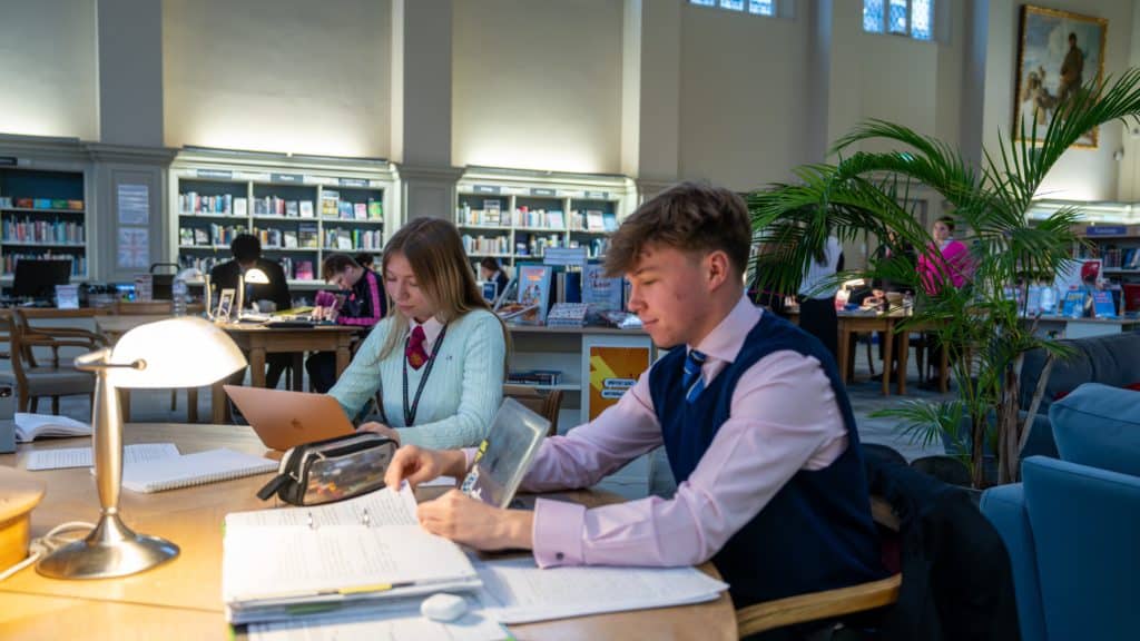 Two students study at a library table with notebooks, pens, and laptops. Bookshelves and other people are visible in the background, with sunlight streaming through large windows.