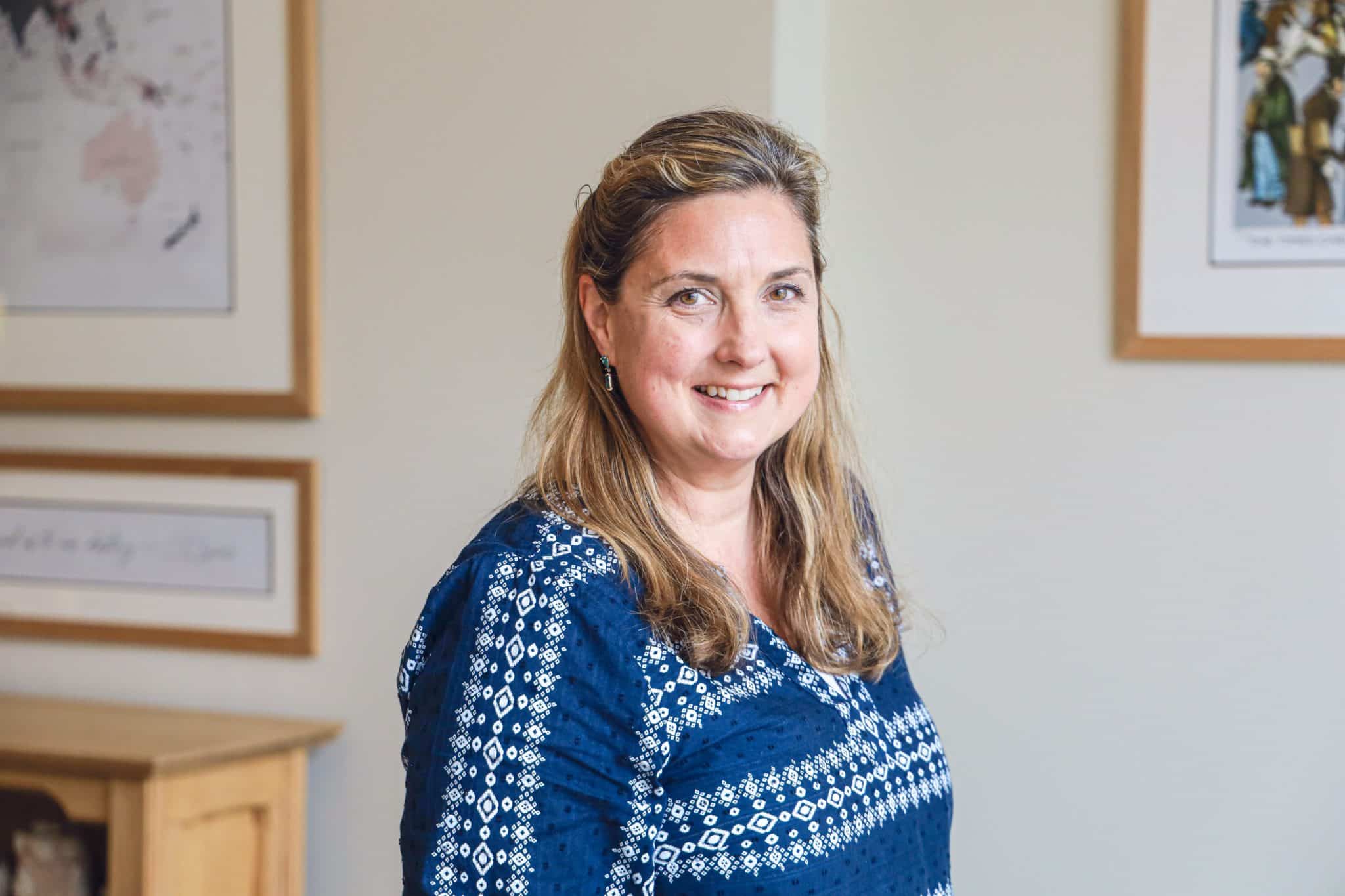A woman with long, light brown hair wearing a blue patterned blouse smiles whilst standing indoors, perhaps ready to discuss Cheltenham College admissions. Framed pictures and light-coloured walls are visible in the background.