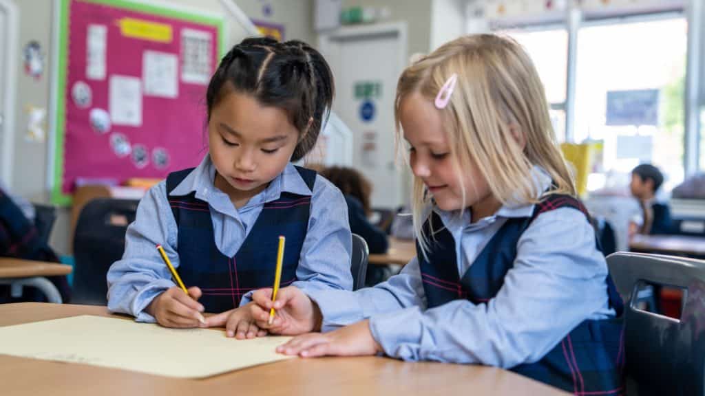 Two young girls in Cheltenham Prep School uniforms sit at a classroom desk, focused on writing with pencils on a large sheet of paper, with a colourful noticeboard and other children in the background.