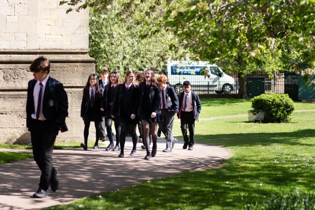 A group of students in school uniforms, perhaps engaged in Oxbridge Preparation, walk along a shaded path in a sunny park, with one student confidently walking ahead of the others. Trees and grass surround them.