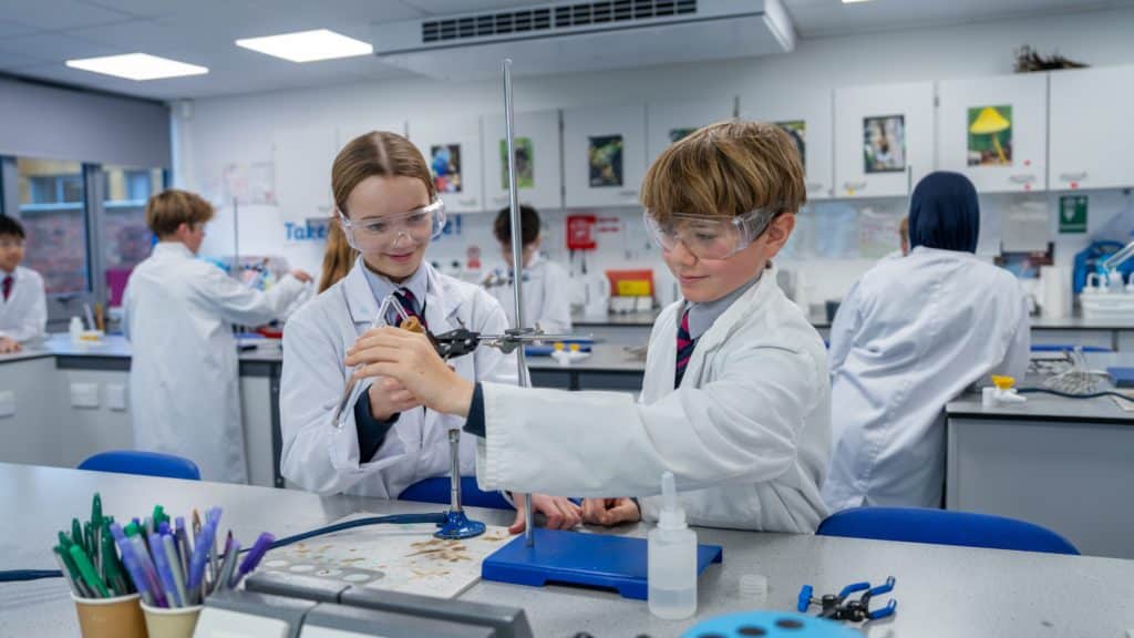 Two students from Cheltenham College, dressed in lab coats and goggles, conduct a science experiment with laboratory equipment at a classroom table, whilst other students work in the background.