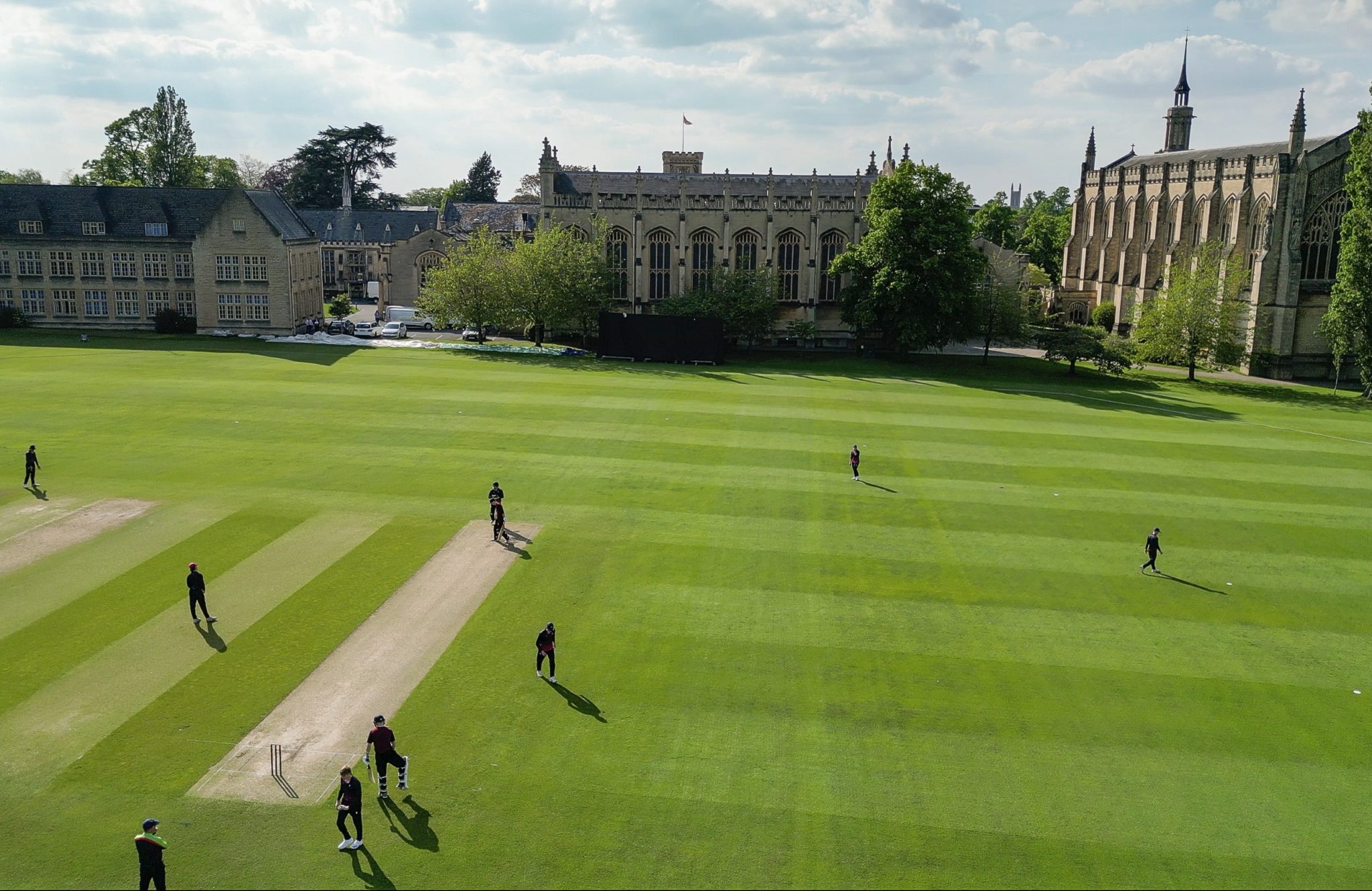 A cricket match is being played on a large green field at Cheltenham College and Prep, with players in dark kit, surrounded by historic stone buildings and trees under a partly cloudy sky.