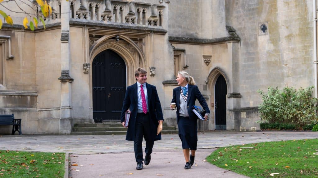 Two people in business attire walk and talk outside a historic stone building with arched doorways, holding folders and coffee cups, surrounded by grass and autumn leaves.