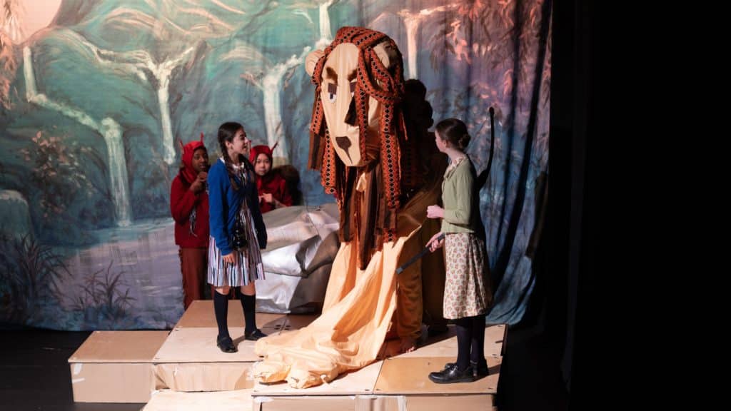 Several children in fancy dress from Cheltenham Prep School perform on stage with a large lion puppet, set against a painted forest backdrop. Two girls stand at the front whilst others, dressed as creatures, stand behind, all engaged in the scene.