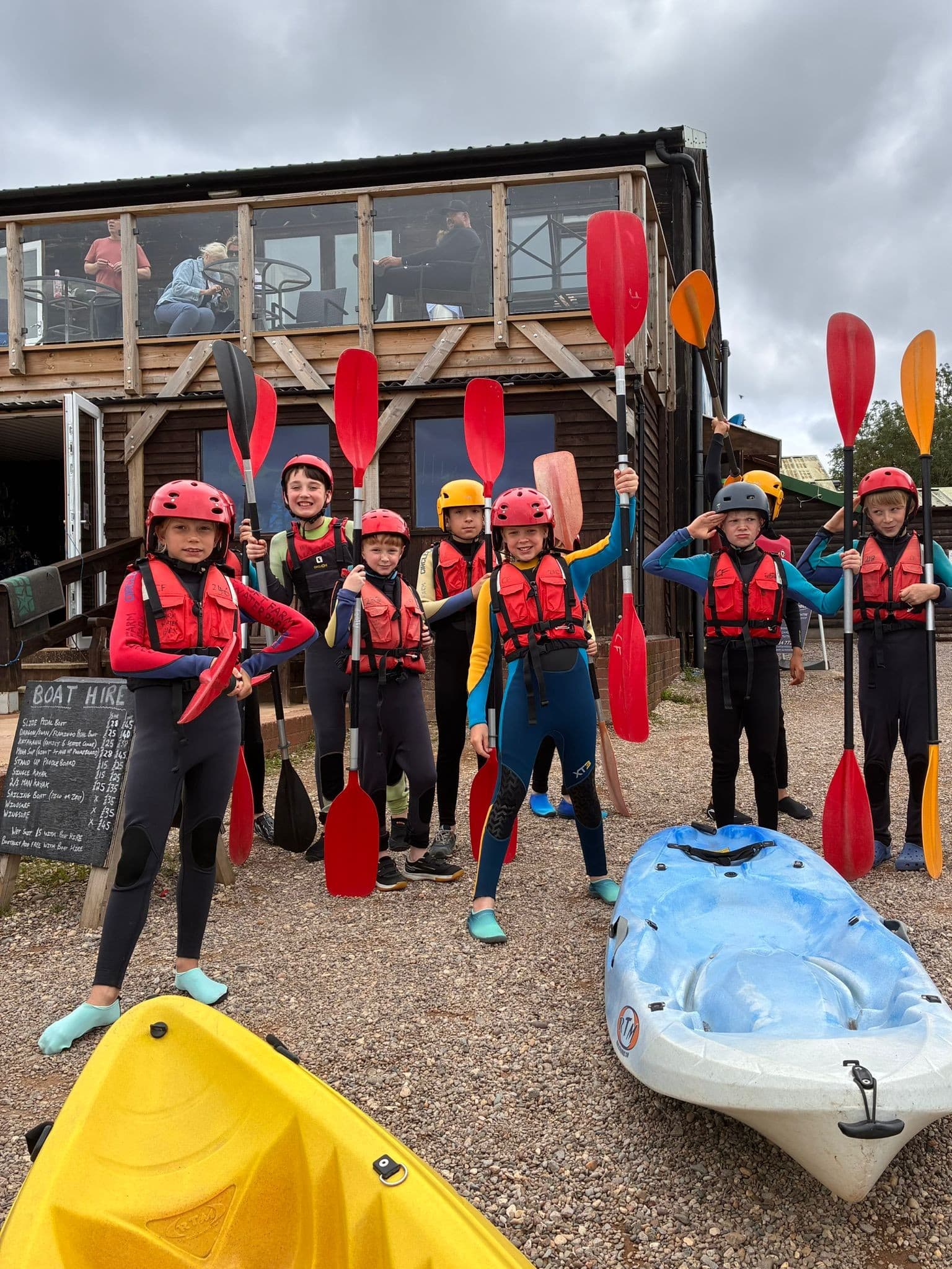 A group of children and adults wearing wetsuits, life jackets, and helmets stand holding kayak paddles in front of a rustic boathouse. Two kayaks are on the ground nearby. The group appears ready for a kayaking activity.
