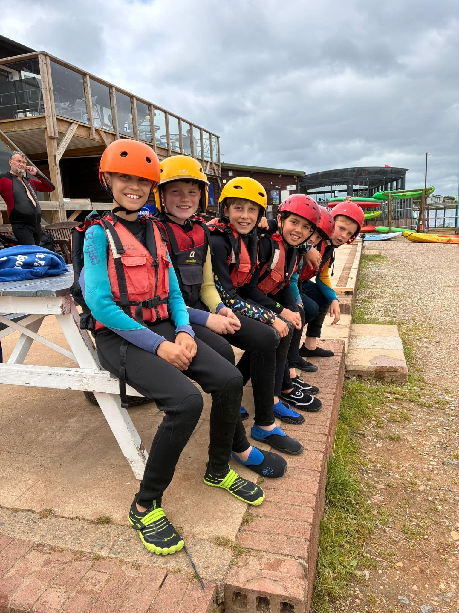 Six children wearing wetsuits, life jackets, and helmets sit on a low brick wall, smiling at the camera. Behind them are wooden buildings, kayaks, and a cloudy sky, suggesting they are at a watersports centre.