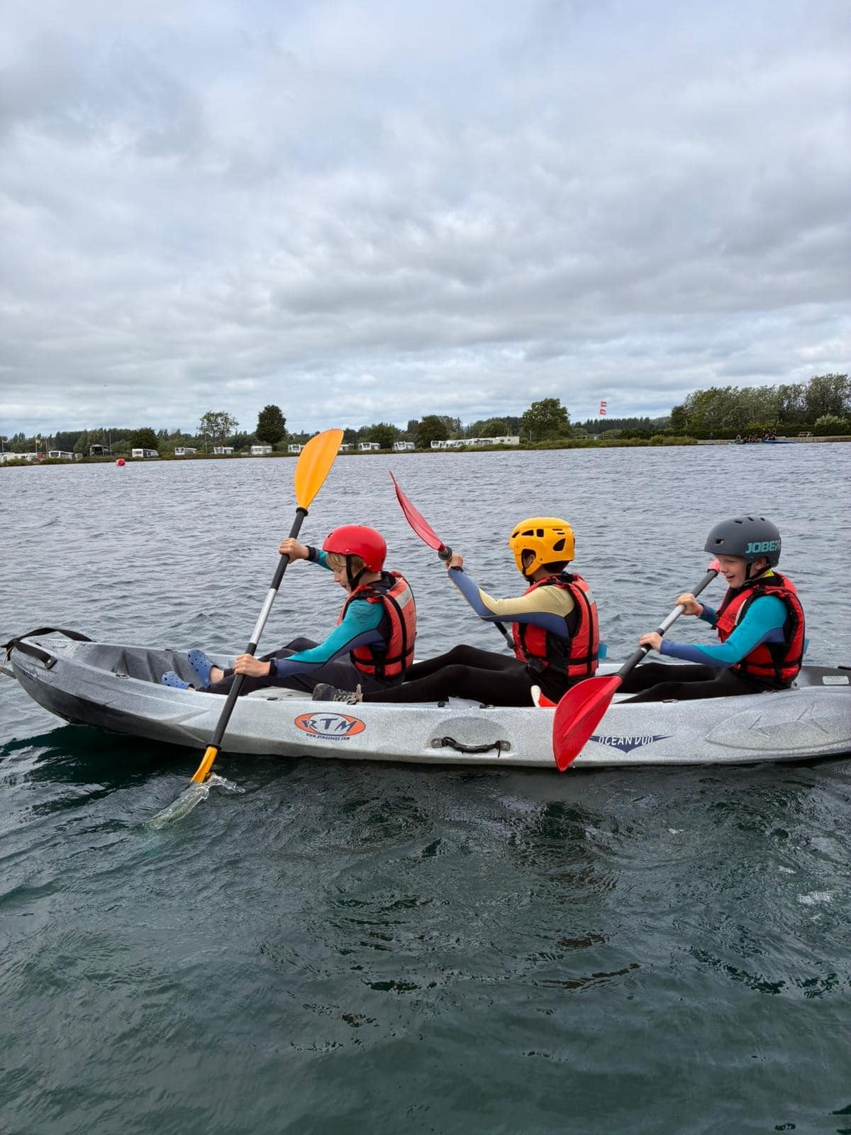 Three people wearing helmets and life jackets paddle a grey inflatable kayak on a lake under a cloudy sky. The water is calm, and trees and buildings are visible in the background.