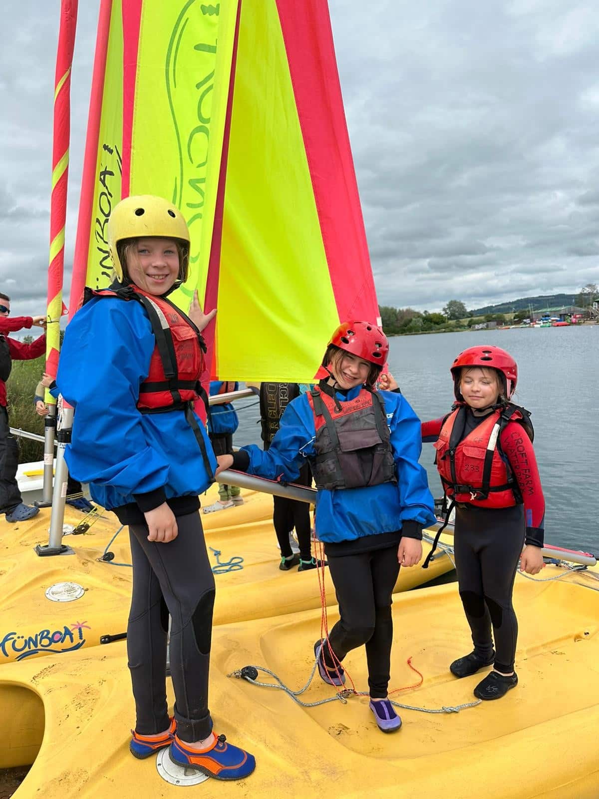 Three children in blue jackets, red life vests, and yellow helmets stand on a yellow sailing boat with bright yellow and red sails on a cloudy day, smiling by the water.