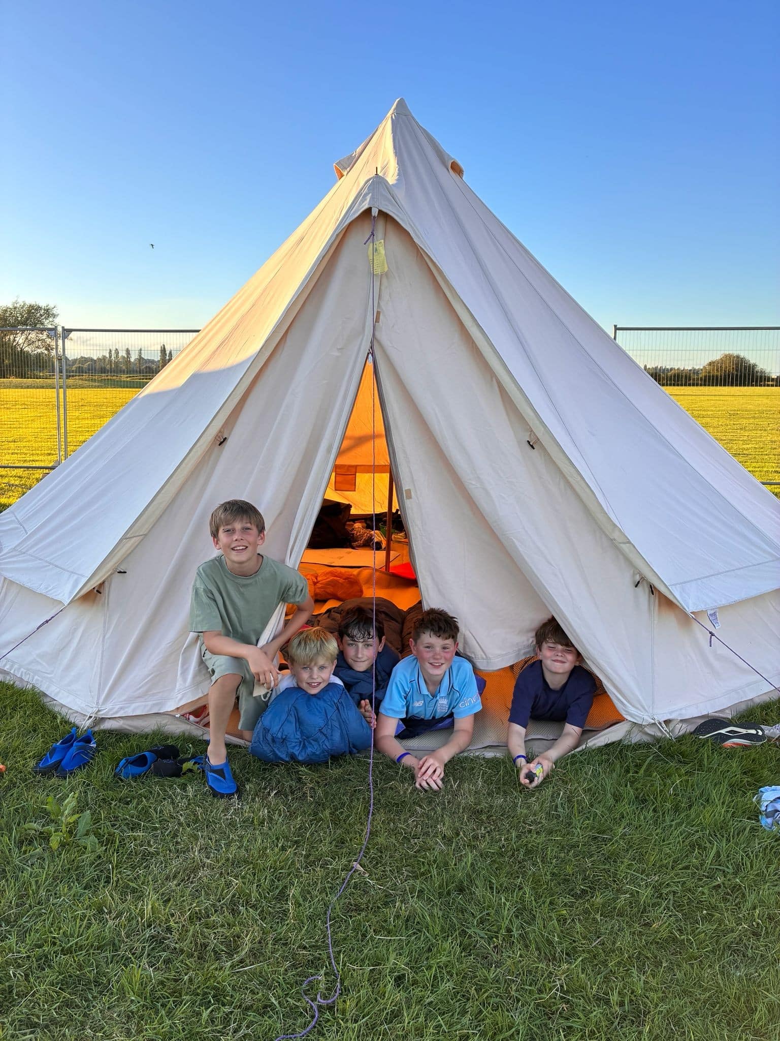 Five boys sit and kneel at the entrance of a cream-coloured canvas tent on green grass, smiling at the camera under a clear blue sky, with sunlight shining inside the tent.