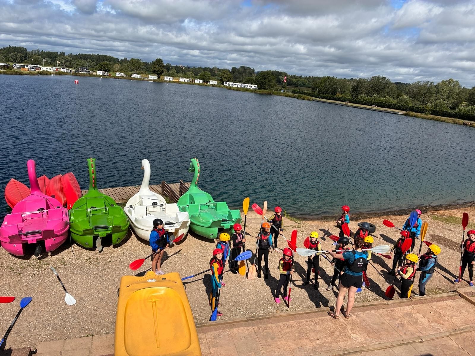 A group of people in life jackets and helmets stand on a lakeshore near colourful pedal boats shaped like swans and ducks, holding paddles, preparing for a water activity under a partly cloudy sky.