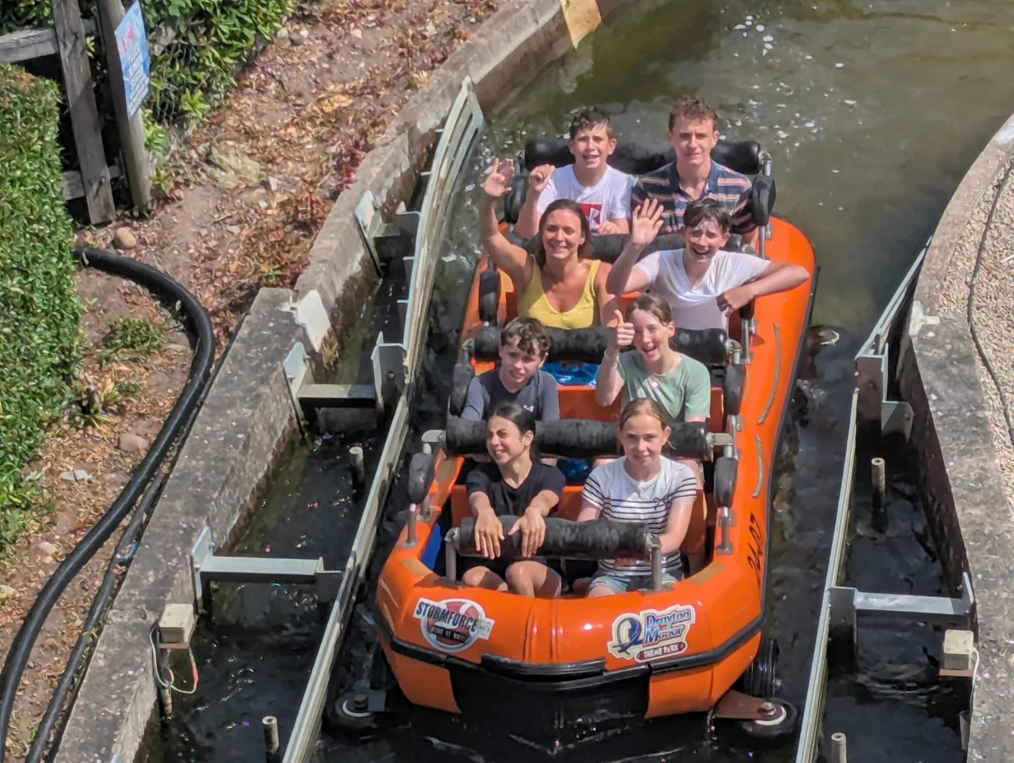 A group of seven people, including children and adults, smile and wave whilst riding a circular orange water raft on a theme park water ride surrounded by concrete walls and water.