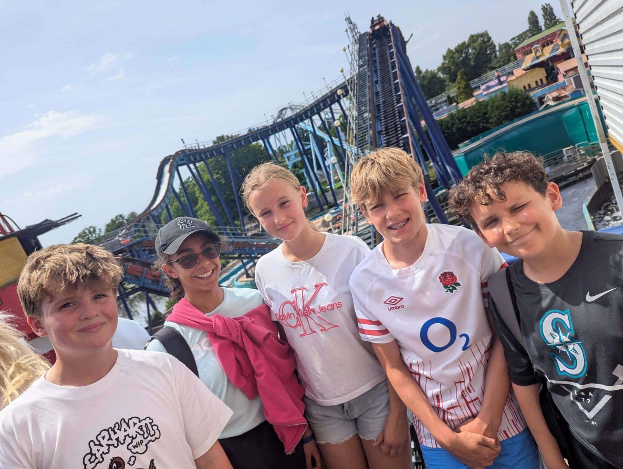 Five smiling children stand together at a theme park with a large rollercoaster in the background. The sky is clear and sunny, and the group appears happy and excited.