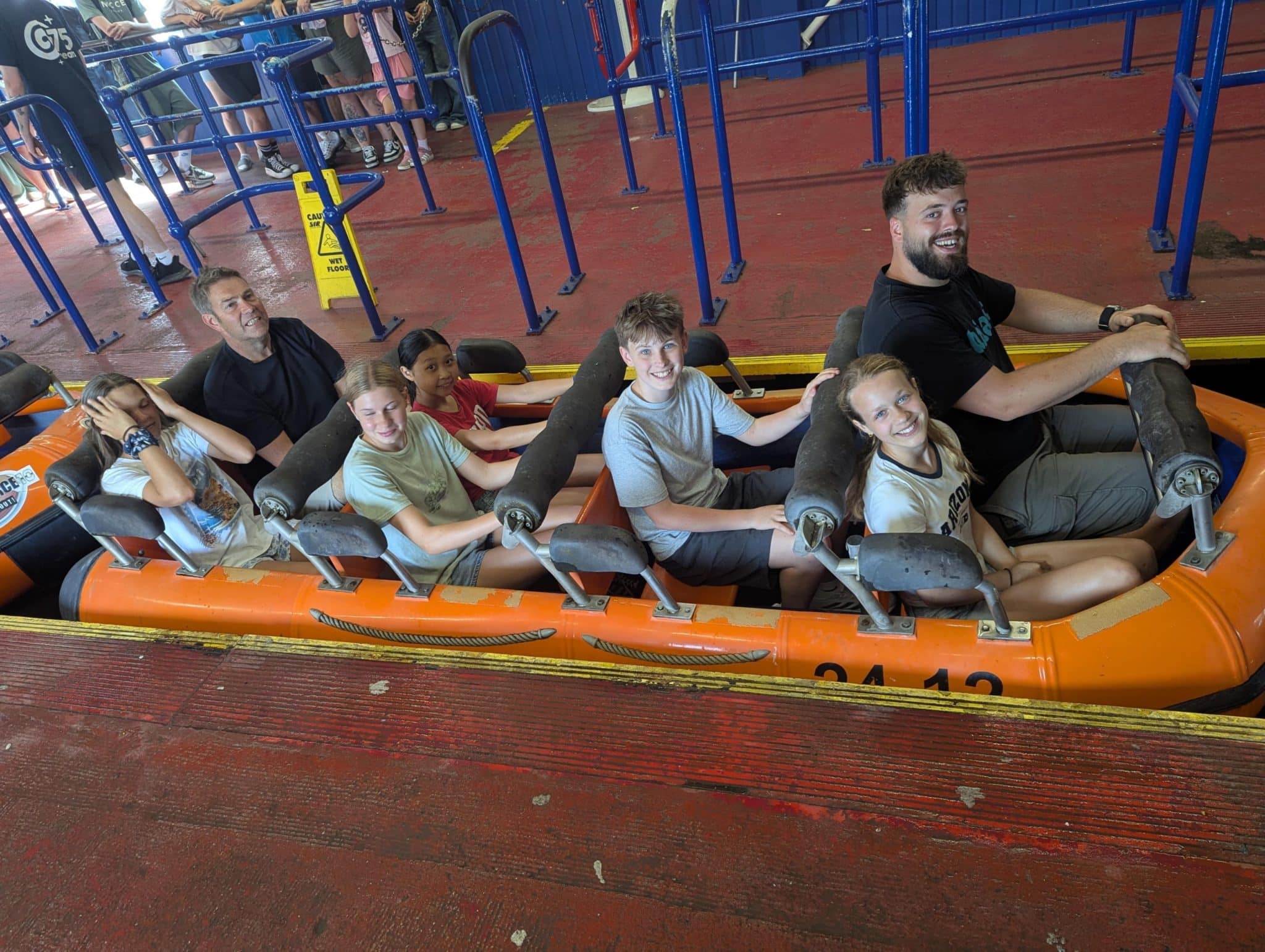 A group of six people, four children and two adults, sit together in an orange amusement park ride, smiling and posing before the ride begins. Blue railings and a red floor are visible around them.