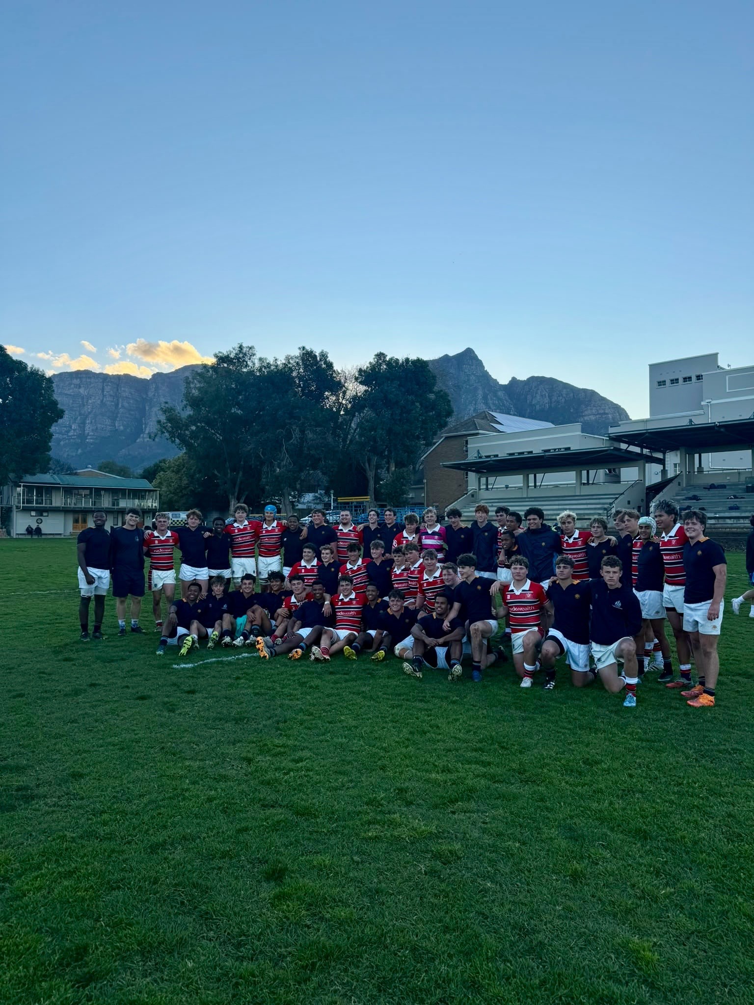 A large group of young rugby players, wearing red and white or dark jerseys, pose together on a grassy pitch in front of school buildings and mountains at sunset.