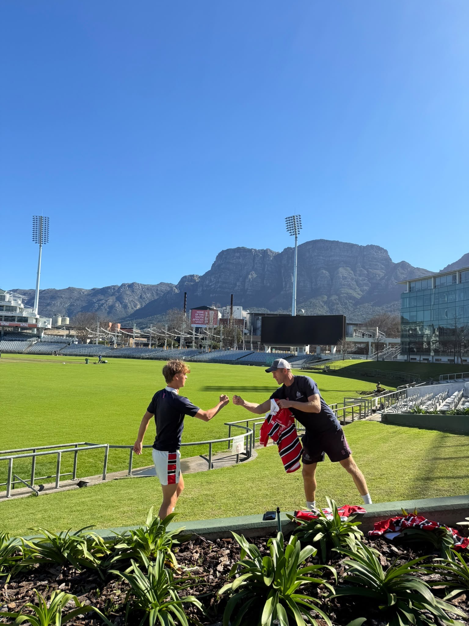 Two people on a sunny stadium pitch fist-bump each other. One holds team shirts. Stadium lights, empty seats, and a mountain range are visible under a clear blue sky. Some flowers and greenery are in the foreground.