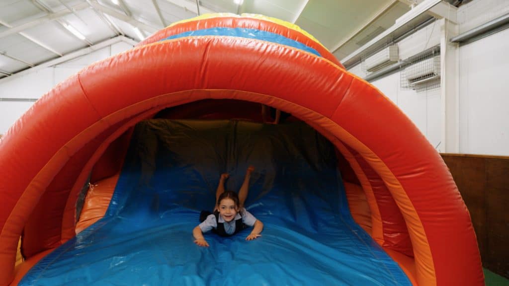 A child slides down a blue and orange inflatable slide inside a large indoor play area, smiling and having fun.