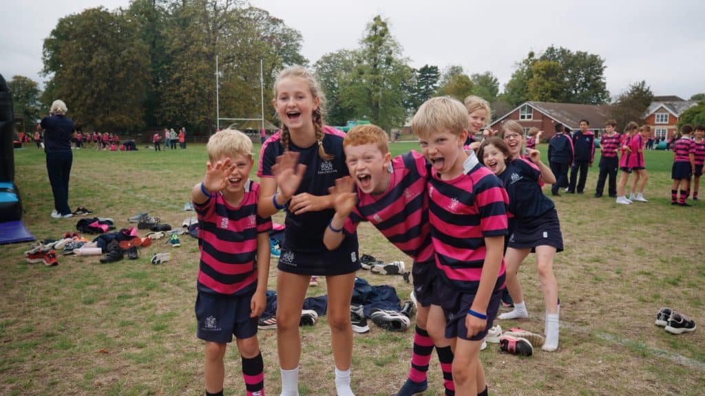 A group of enthusiastic children in matching sports kits smile and wave at the camera on a grassy field, with more children and trees in the background.